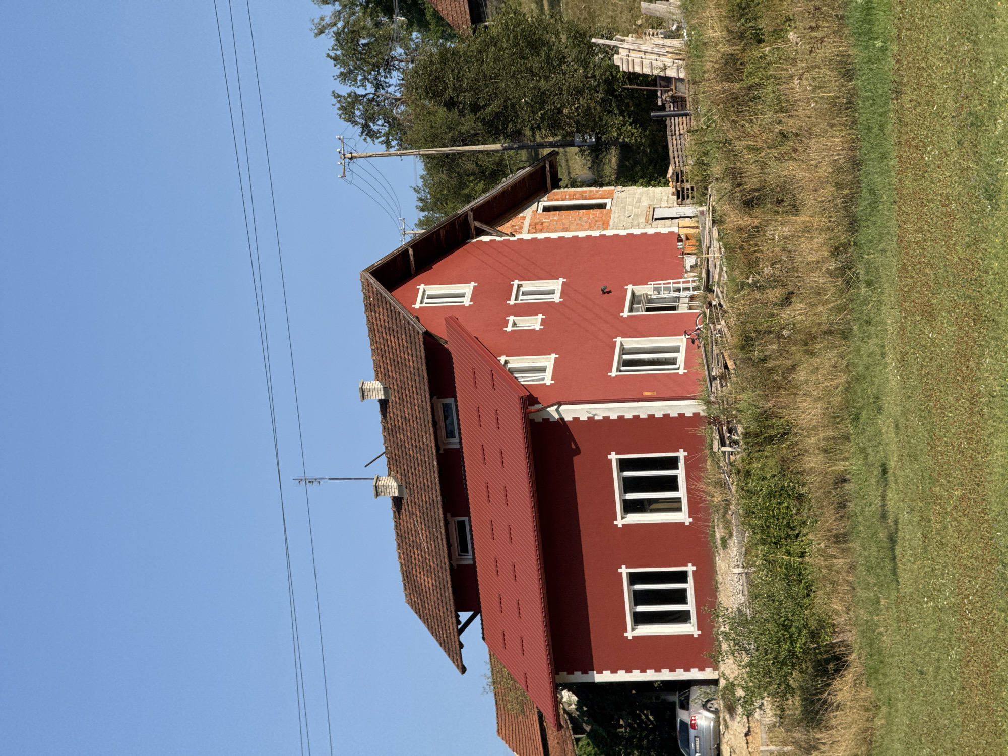 The image depicts a two-story house with a red facade and a tiled roof. The house is situated in a rural area with a clear blue sky overhead. In front of the house, there is a field of dry grass and some green vegetation. To the left of the house, a silver car is parked. In the background, there are trees and a wooden utility pole with power lines. The house appears to be under construction or renovation, as indicated by exposed brickwork on one side and stacks of wood and construction materials nearby. The scene suggests a residential property in a somewhat remote location. The location is identified as Cajetina Municipality, Serbia.