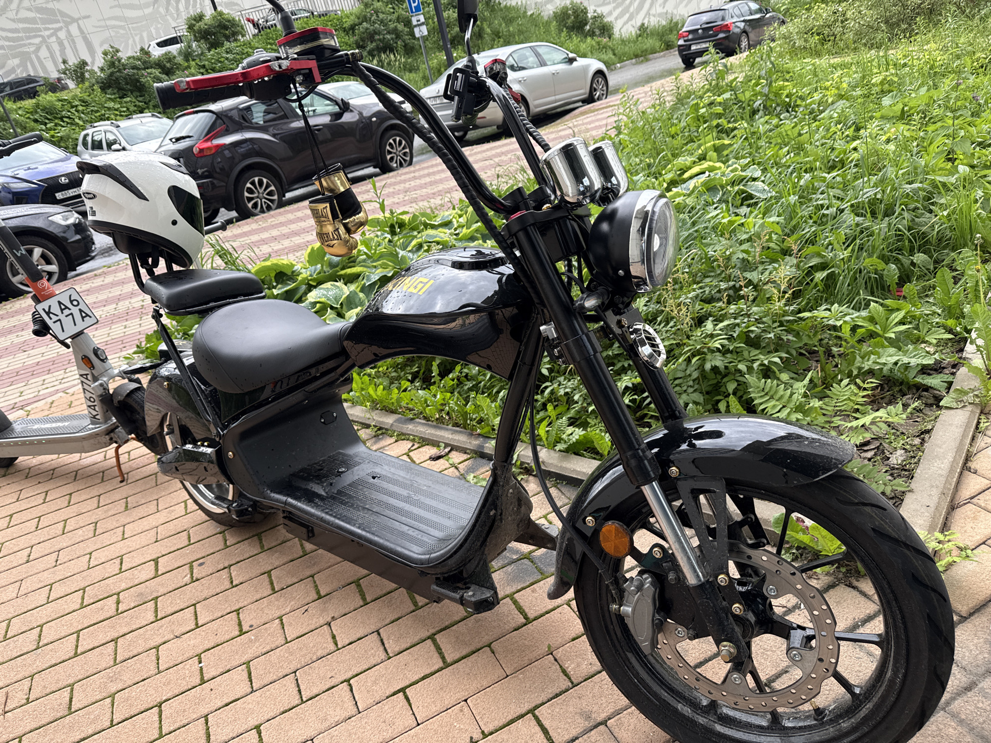 The image shows a black electric scooter parked on a brick sidewalk. The scooter has a white helmet on the seat and a pair of gold boxing gloves hanging from the handlebars. In the background, there are several cars parked on a street and a building with a patterned facade. The vegetation is lush and green, suggesting a temperate climate. The license plate on the scooter reads "KA677A". Based on the presence of the license plate format and the style of the cars, the location appears to be Moscow, Russia.