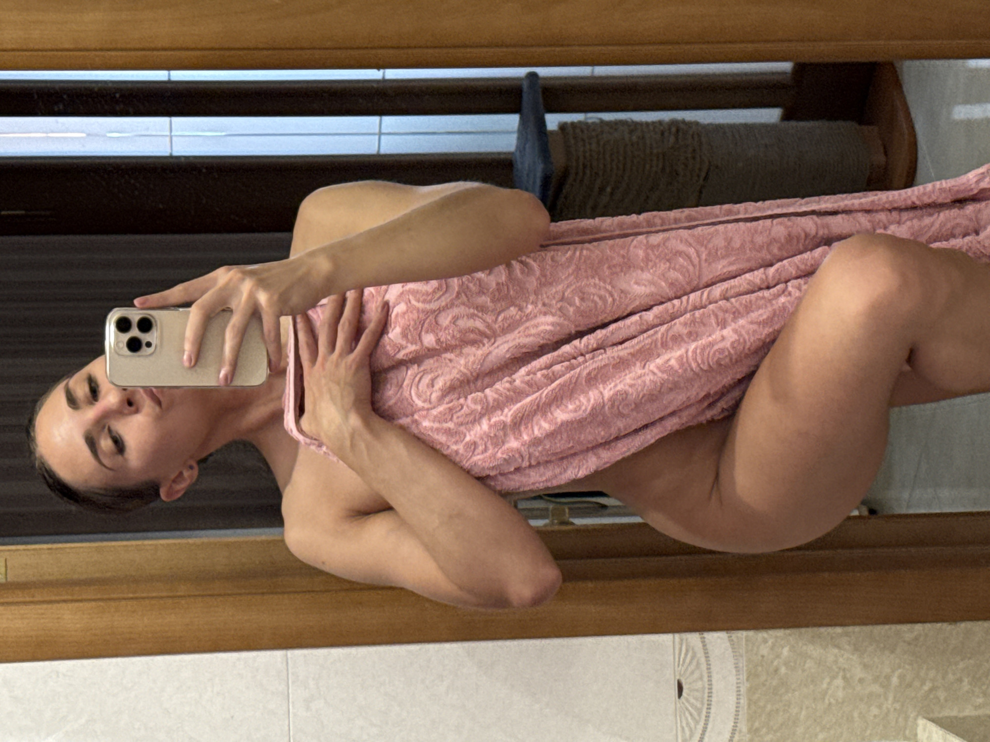 The image displays a mirror selfie of a person, likely female, captured in an indoor setting. The individual is positioned horizontally in the frame, with their head to the left and legs to the right, and is partially draped in a light pink towel or robe featuring a subtle, embossed pattern.
The person is holding a light-colored smartphone, equipped with a clear case and a triple camera array, in their right hand. Their left hand also rests on the pink towel. The individual's face is visible, characterized by dark eyebrows, prominent eyelashes, and a slightly pouting expression. Their dark hair appears to be pulled back.
Reflected in the mirror, forming the background, are several elements. A prominent wooden frame runs horizontally across the upper part of the reflection. Above this frame, there are faint horizontal white lines against a dark background, possibly reflections of windows or internal lighting. Below the person's body, particularly near their legs, a large cylindrical object wrapped in coarse, light brown rope or fabric is visible, resembling a pet scratching post or a decorative item. A dark blue or grey object, possibly fabric, is also seen near this cylindrical item. The very bottom of the reflection reveals a white tiled surface or countertop, which includes a tile with a subtle, circular embossed pattern.
The setting appears to be a private room, such as a bathroom or dressing area, indicated by the mirror, towel, and tiled surfaces. The time of day is not discernible due to artificial lighting, and there is no visible text in the image.