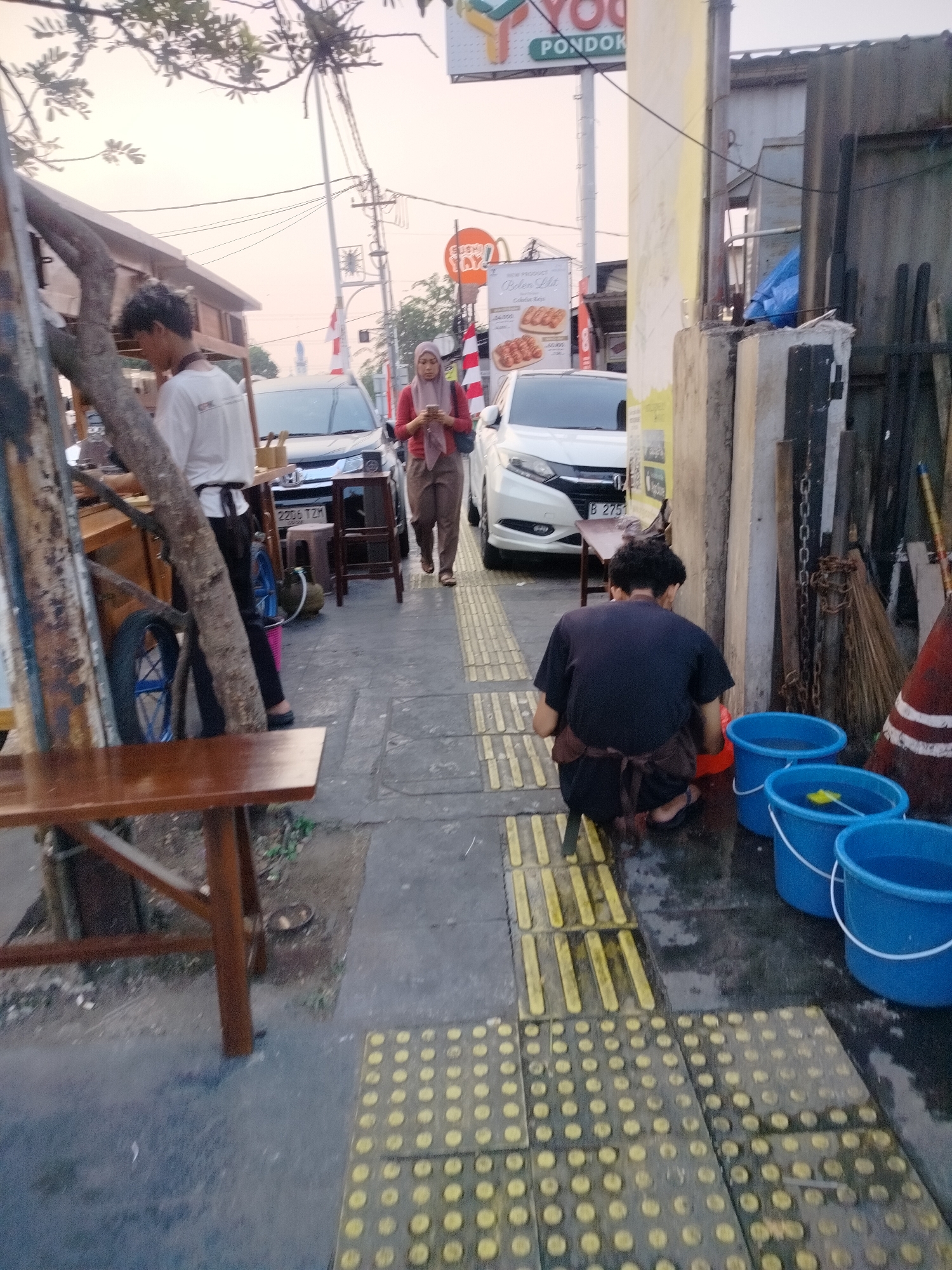 In East Jakarta, Indonesia, an outdoor scene unfolds along a sidewalk adjacent to a street. The time appears to be late afternoon or early evening, with the sky showing a soft, muted light.
A person wearing a white shirt and black pants stands behind a wooden food cart, which has a tree trunk growing vertically through its structure. The cart appears to be set up for business, with various items on display. Further down the sidewalk, a woman in a maroon hijab and beige pants walks while looking at her phone.
On the right side of the image, a man wearing a dark blue t-shirt and a brown apron is crouched down, seemingly working near several bright blue buckets of water. A broom leans against a building, and various wooden planks and metal structures are visible, suggesting a utilitarian or working area.
In the background, two cars are parked on the street. One is a white sedan, and the other appears to be a darker vehicle. A sign with the text "YOG PONDOK" is visible in the upper portion of the frame, indicating the area or a business. The sidewalk itself is marked with tactile paving, designed to assist visually impaired individuals, in a pattern of raised yellow dots and bars. The ground is wet in places, possibly from recent rain or cleaning activities.