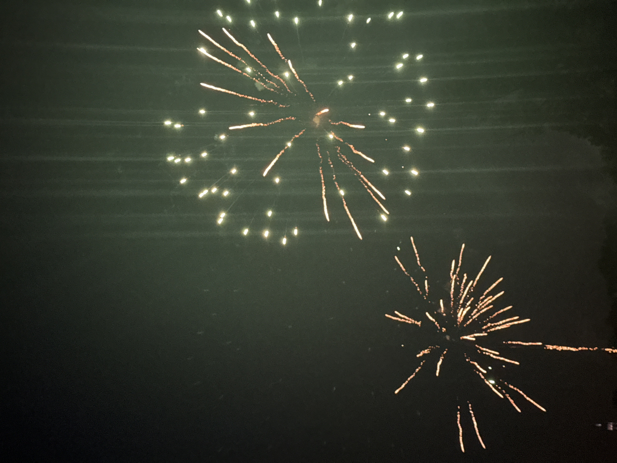 The image depicts a nighttime sky with fireworks exploding. There are two main bursts of fireworks visible. The one on the left is a smaller, more scattered display of golden-orange streaks radiating outwards from a central point, with a single vertical trail below it. The larger firework on the right is a more circular burst of numerous small, bright white and yellow lights, appearing more diffuse. The sky is dark green with some faint vertical light streaks, possibly from other fireworks or atmospheric conditions. There are some subtle speckles across the image, suggesting it may be raining or dusty. In the lower left corner, a faint silhouette of a tall, slender structure, possibly a building or monument, can be discerned against the dark horizon. The lower edge of the image shows a dark, indistinct silhouette of trees or foliage. The overall scene suggests a fireworks display taking place at night.