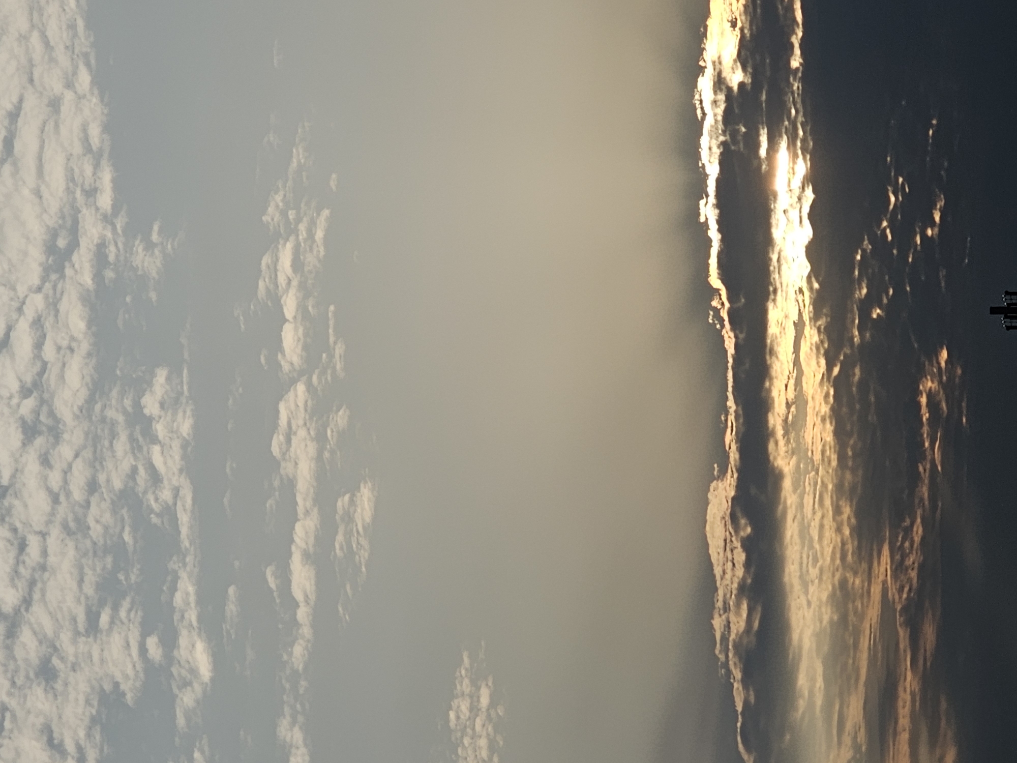 The image depicts a sky with layered clouds, some illuminated by the sun breaking through. The upper portion of the sky is a pale blue with scattered, fluffy white clouds. Towards the horizon, darker, denser clouds are visible, with the sun creating a bright glow and rays of light piercing through them. The overall atmosphere suggests a sunset or sunrise. A small portion of what appears to be a dark, man-made structure, possibly a tower, is visible at the very bottom of the image, silhouetted against the horizon. The image does not contain any people or specific identifiable landmarks of Khimki, Russia.