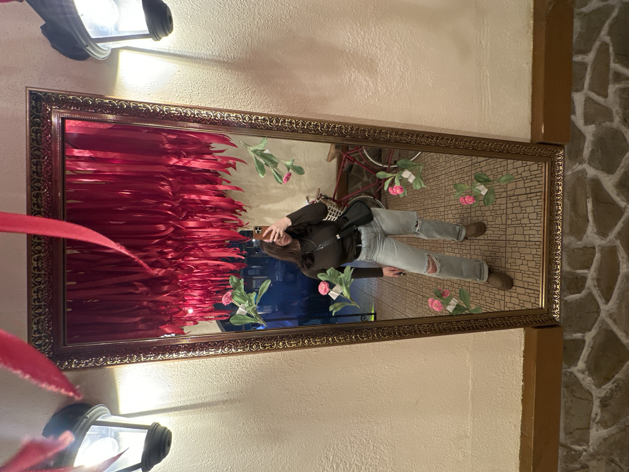 The image depicts an indoor scene in Chaikovsky, Russia, centered around a tall, rectangular mirror with an ornate gold-colored frame. A young adult female is reflected in the mirror, holding a light-colored smartphone with her right hand to take a selfie; her face is partially obscured. She is dressed in a dark brown long-sleeved top, light-wash ripped jeans, and dark brown shoes, with a small black shoulder bag worn across her body.
Within the mirror's reflection, above the female's head, numerous red ribbons hang vertically, creating a dense curtain-like effect. To her left in the reflection, a vintage-style red bicycle with a front basket is visible. The reflected floor shows a brown, patterned tile design. Several artificial pink flowers with green leaves are attached directly to the surface of the mirror glass.
Outside the mirror frame, a textured, light-colored wall serves as the background. Two black, lantern-style wall sconces are mounted on this wall, one on each side of the mirror, emitting bright light. Additional red ribbons are visible hanging from above, outside the mirror's top edge. A dark wooden baseboard runs along the bottom of the wall, and the floor in the immediate foreground consists of irregular, earthy-toned stone or tile patterns.