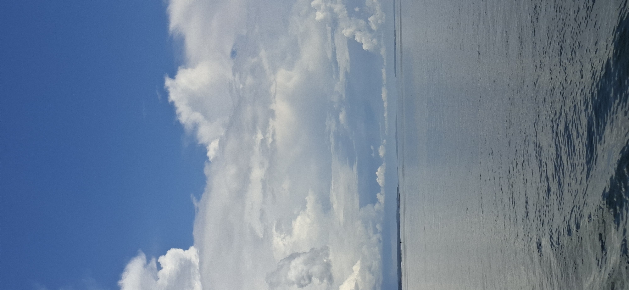 This is a vertical, eye-level, full-shot photograph of a vast expanse of water under a dynamic sky. The upper portion of the image is dominated by a bright blue sky filled with a mixture of large, puffy white cumulus clouds and more diffused, greyish clouds. The clouds are particularly dense and textured in the middle section of the frame, creating a dramatic contrast with the clear blue sky above.
The lower two-thirds of the image depict a calm, reflective body of water. The surface of the water is smooth with subtle ripples, creating a mirror-like effect that reflects the colors of the sky. The water transitions from a lighter, almost greyish hue near the horizon to a darker, more saturated blue towards the foreground, where the texture of small waves becomes more apparent.
In the distance, along the horizon, there is a faint, elongated landmass, suggesting a coastline or a chain of islands. The land is low-lying and appears densely vegetated, with a few dark spots that could be trees or buildings. A very small, dark vertical marker, possibly a buoy or a navigational aid, is visible on the water near the left side of the horizon.
The scene evokes a sense of tranquility and openness, typical of a coastal or large body of water environment. Given the context of Helsinki, Finland, this image could plausibly be a view of the Baltic Sea from one of its many islands, peninsulas, or a ferry. The presence of islands in the distance further supports this interpretation, as the Helsinki archipelago is extensive. The overall atmosphere is one of natural beauty and a serene day by the water.