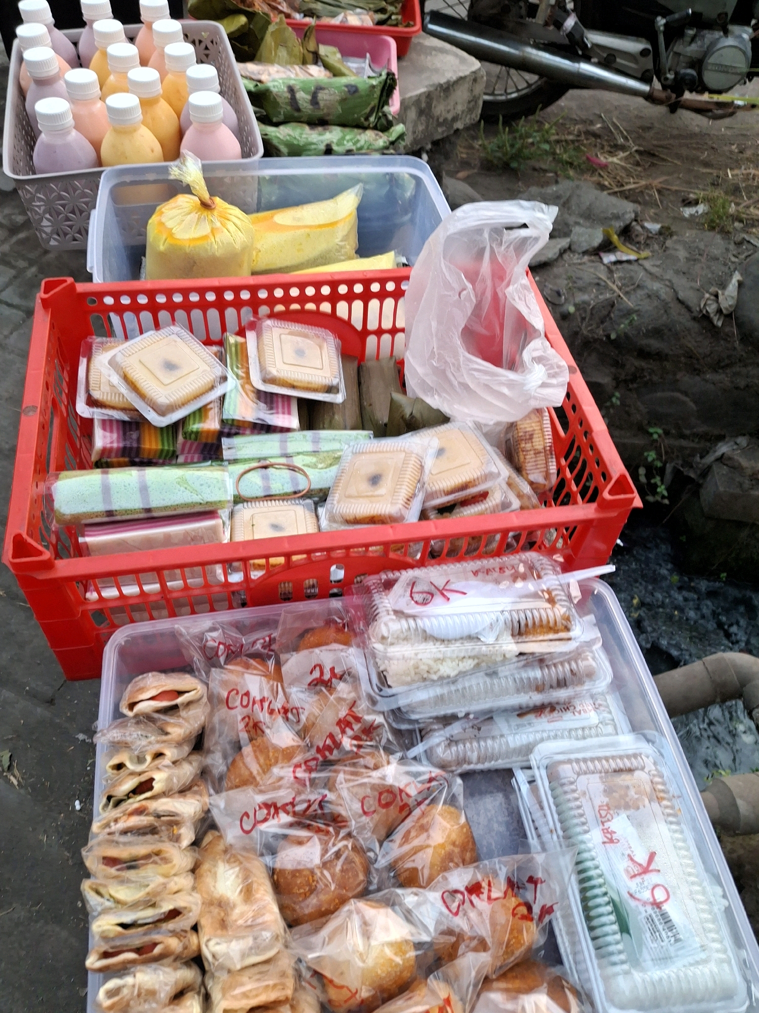 The image depicts an outdoor market stall, likely in Lumajang, Indonesia, displaying various food items for sale. In the foreground, a clear plastic container holds an assortment of pastries and bread, individually wrapped in clear plastic. Some of these items have red handwritten labels, including "6K" and "Coklat". To the left of this container, a stack of small, folded savory pastries, appearing to contain a red filling, are also individually wrapped.
Behind this, a red plastic crate is filled with pre-packaged desserts. These include rectangular cakes in clear plastic containers, some with a layered appearance in various colors, and others that are a solid yellow or brown hue. One container holds what appears to be a yellow cake or jelly, wrapped in a yellow plastic bag tied with a rubber band. To the left of the red crate, a white plastic basket contains several small, clear bottles with white caps, filled with what appears to be colored liquid, likely yogurt drinks in shades of pink and yellow.
In the background, there are other food items wrapped in green leaves and some plastic packaging. A motorcycle is partially visible, with its exhaust pipe resting on the ground, suggesting the stall may be set up alongside a road or pathway. The ground surface appears to be a mix of dirt, gravel, and some debris, with hints of vegetation. The overall scene suggests a typical street-side food vending operation.