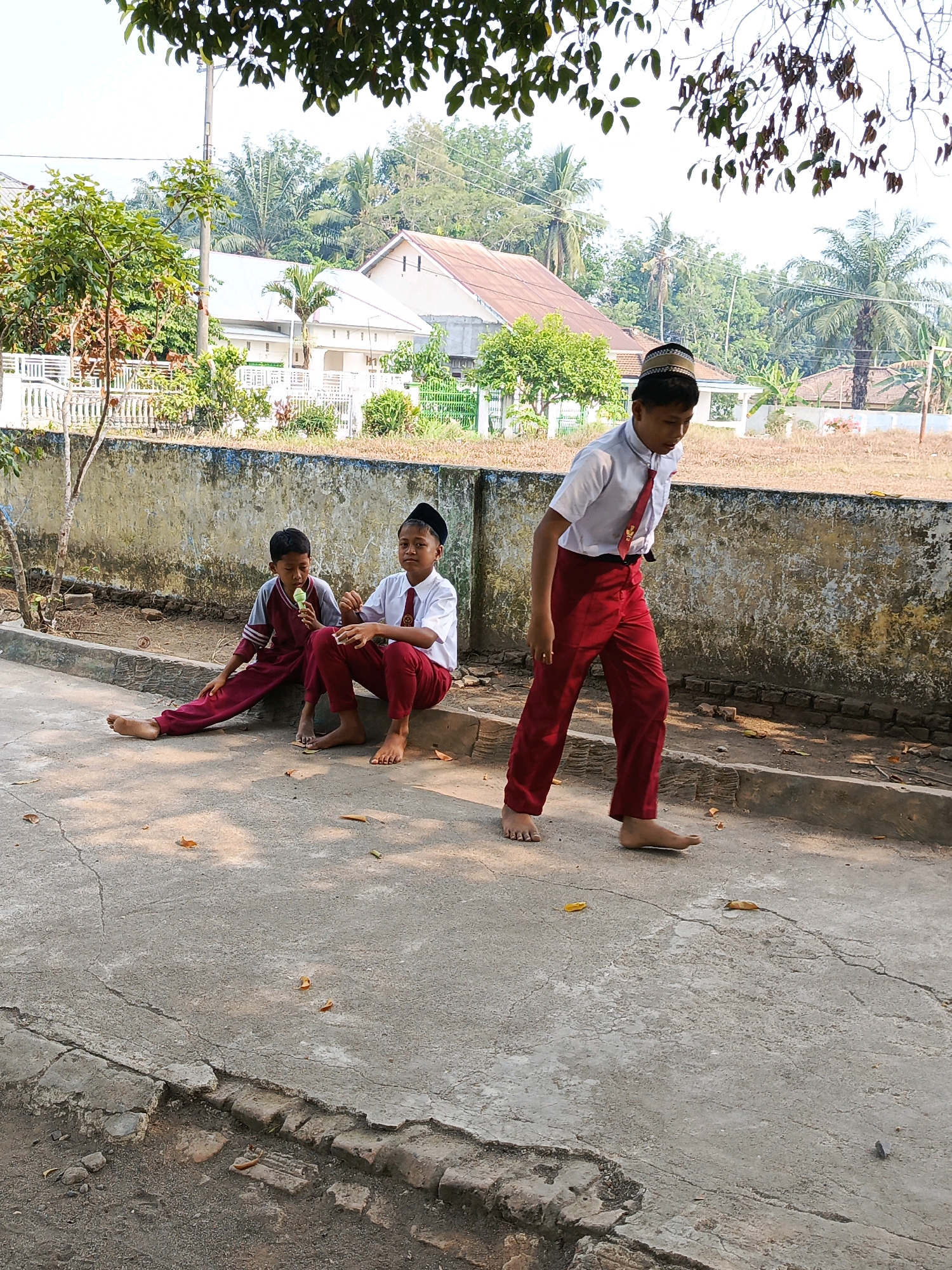 In Purwakarya, Indonesia, three school-aged boys, dressed in red pants and white shirts, are present. Two boys sit on a concrete curb, one holding an ice cream cone, while the third boy stands and walks away from the camera. The background features a low, textured wall, houses with peaked roofs, and lush green trees. The ground is paved with concrete, showing cracks and scattered leaves. The lighting suggests it is daytime, with shadows cast on the ground.