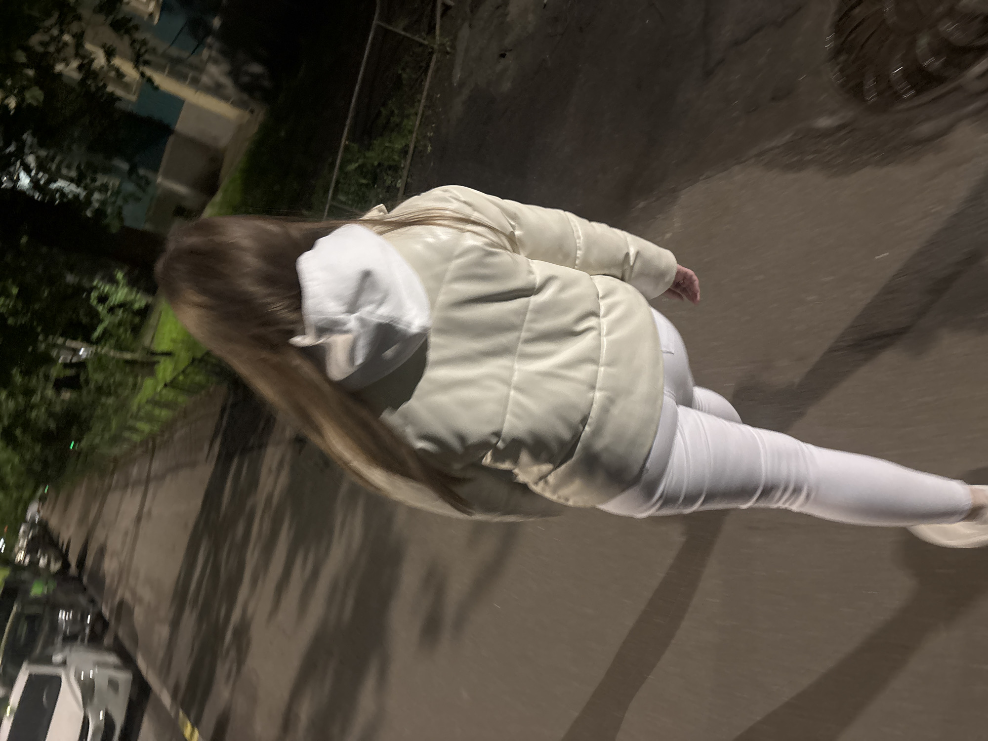 A full shot depicts a fair-skinned individual walking outdoors at night, clad in all white attire: a hooded jacket, pants, and shoes. The ground beneath appears to be an asphalt surface. In the vicinity are trees and buildings, visible on the left side of the shot. The lighting conditions suggest it is nighttime.