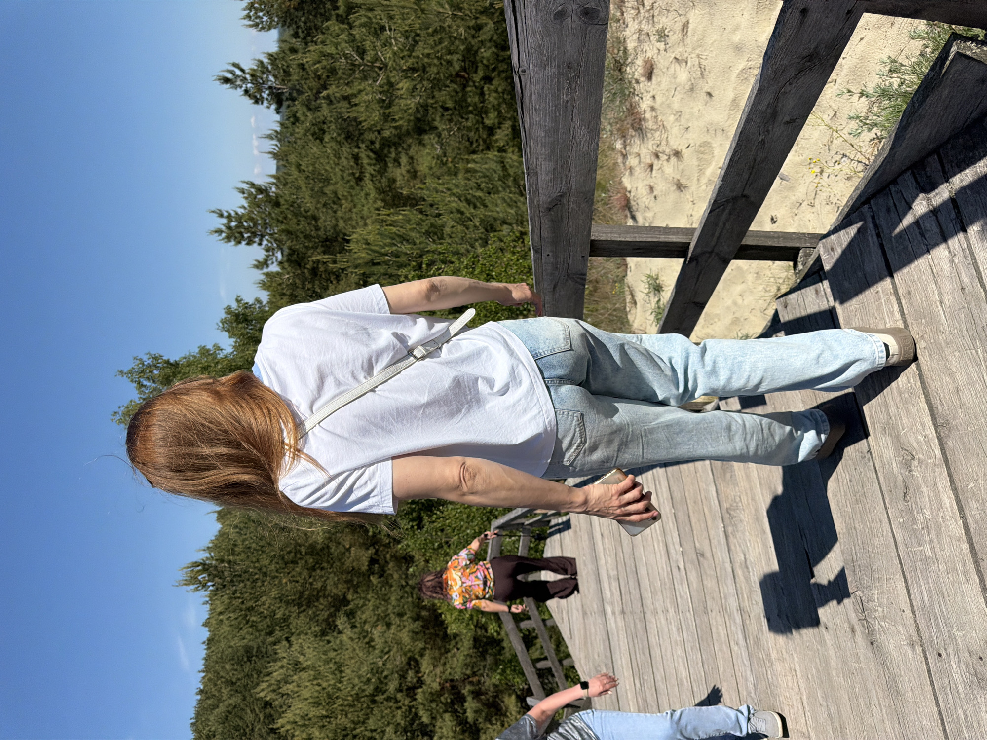 In Morskoye, Russia, a group of individuals ascend a wooden walkway situated amidst sandy dunes and dense coniferous forest. The sky is clear and blue. The foreground features a woman from the rear, with long auburn hair, wearing a white t-shirt and light blue jeans. She holds a mobile phone in her right hand. To her left, another person in light blue jeans and a grey top is partially visible. Further up the walkway, a third person, viewed from behind, is dressed in a colorful patterned shirt and dark brown flared pants. The walkway is constructed of weathered wooden planks and features a dark wooden railing. The environment suggests a natural, outdoor setting.