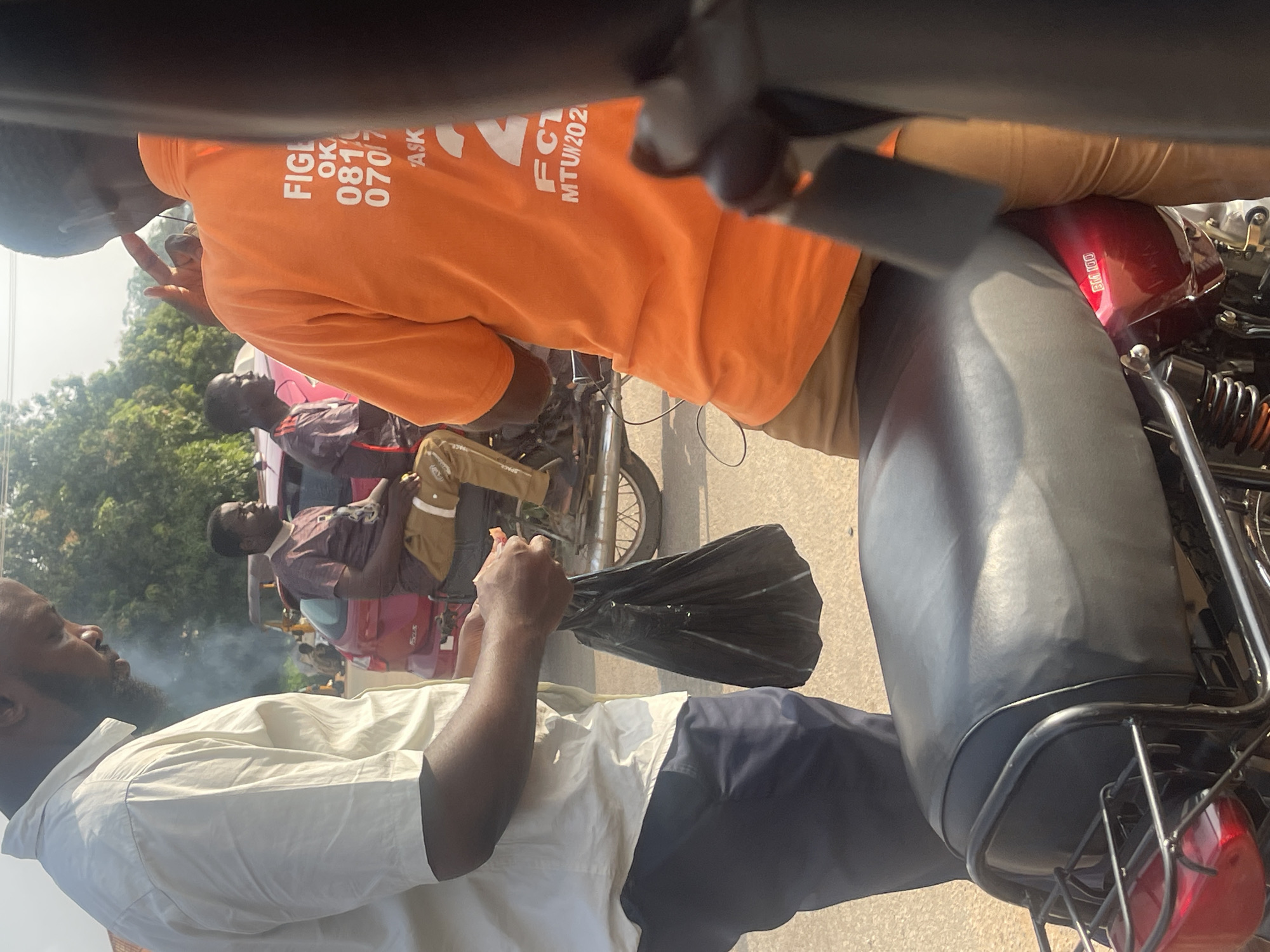 The image depicts a street scene in Galadimawa Village, Nigeria, with a focus on a group of individuals and motorcycles. In the foreground, a person wearing an orange t-shirt with white lettering, including "FIGL" and a phone number starting with "081," is partially visible from the back. They are standing next to a motorcycle with a black seat and a metallic frame. To the left, another individual in a light-colored shirt and dark pants holds a black plastic bag. In the background, two men are seated on a motorcycle. One is wearing a maroon t-shirt and brown pants, while the other is in a similar maroon t-shirt. A red vehicle is parked behind them. The environment appears to be a roadside with trees and other vehicles visible in the distance, suggesting a typical urban or semi-urban setting. Smoke is rising in the background.