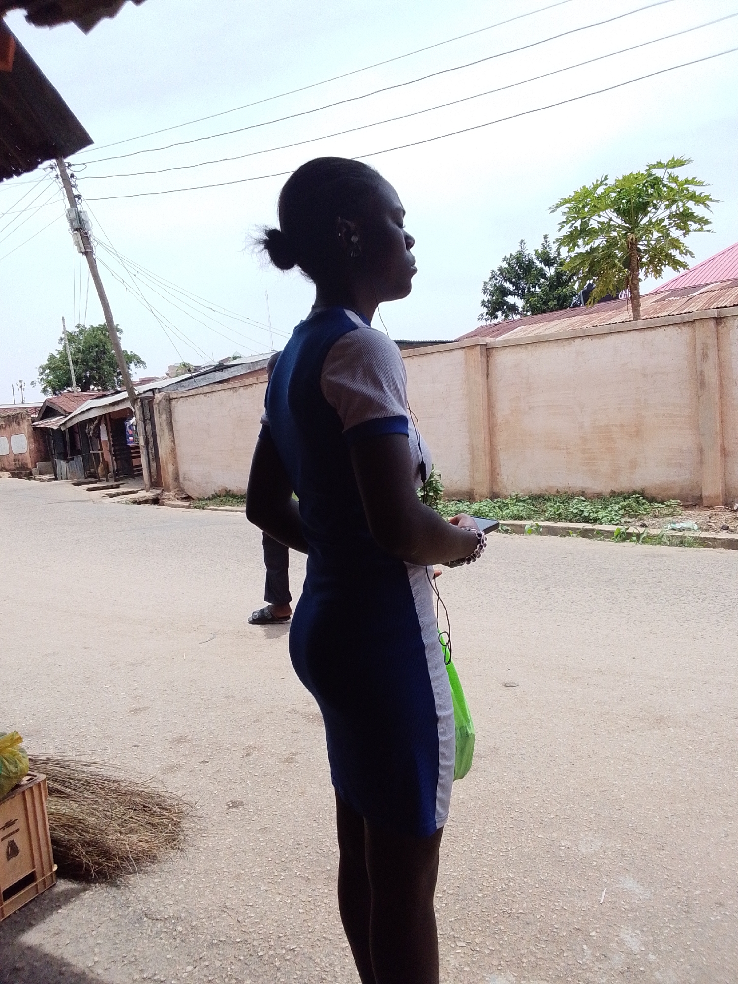 A medium outdoor shot depicts a woman in a blue and white dress standing on a paved road in Jos, Nigeria. She is positioned in the foreground, viewed from the side, with her head turned to the right, and appears to be listening to headphones. She holds a smartphone and has a green bag hanging by her side.
In the background, a concrete wall lines the opposite side of the road, with vegetation visible at its base. A tree is visible above the wall. On the left, there are open-front shops or stalls, with a person standing near one. Overhead power lines stretch across the sky. A crate, some broom, and another person in sandals are also visible. The overall scene is a daytime street view, with a cloudy sky.