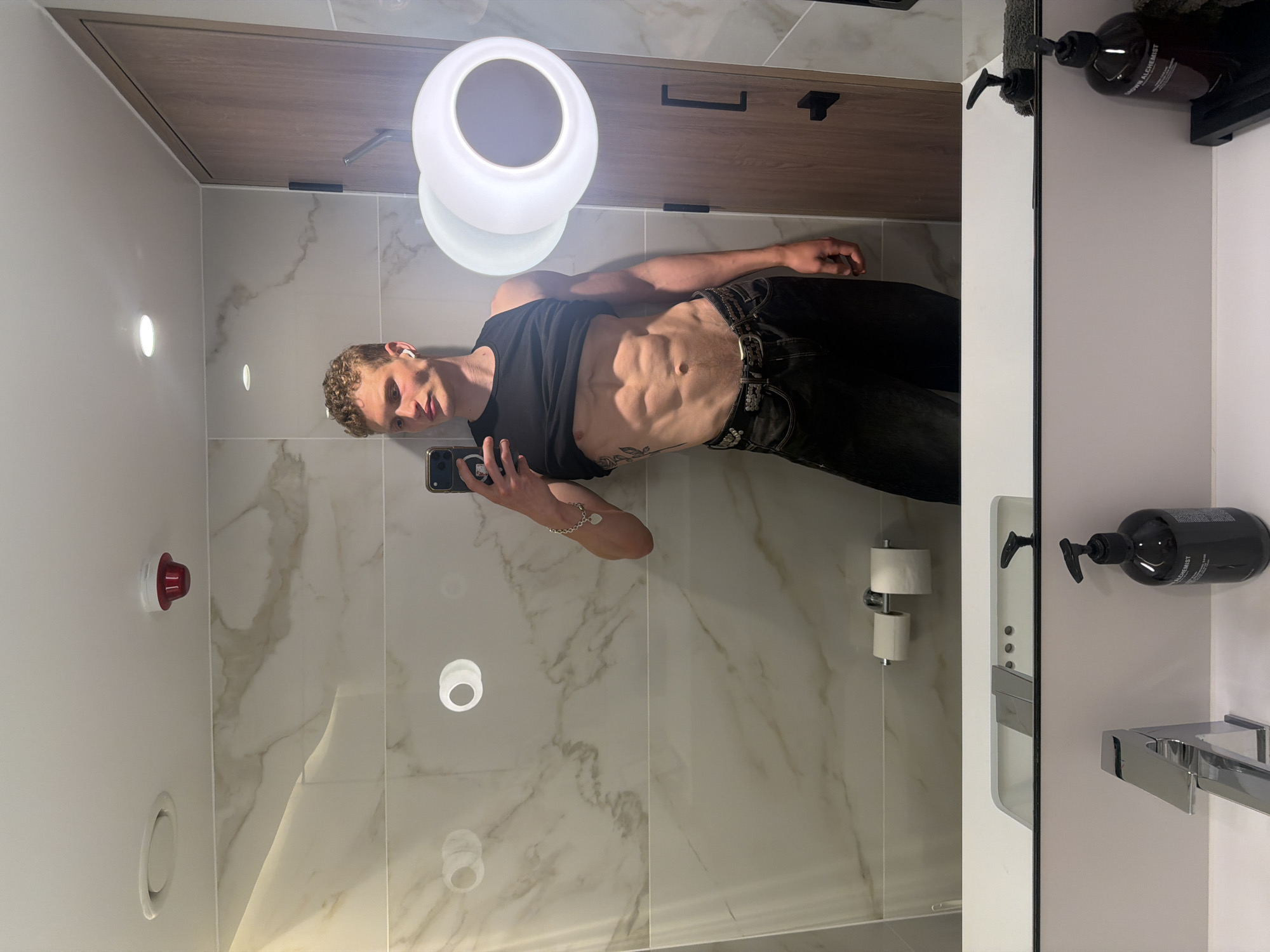 A man is standing in a bathroom, taking a selfie in the mirror. He is wearing a black tank top and black jeans. His abs are visible. There is a light fixture above him. The bathroom has white marble walls. There is a soap dispenser and a towel rack on the right side of the mirror. A roll of toilet paper is attached to the wall. The location cannot be determined based on the information available in the image.