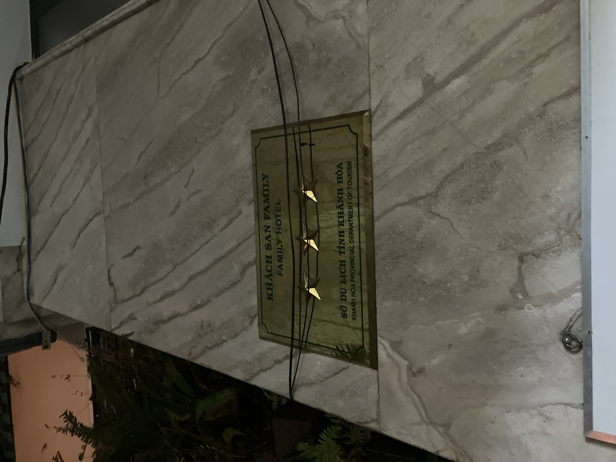 The image shows a sign for "KHÁCH SẠN FAMILY HOTEL" on a marble-tiled wall. Below the hotel name, there is another line of text that reads "SỞ DU LỊCH TỈNH KHÁNH HÒA" and "KHANH HOA PROVINCIAL DEPARTMENT OF TOURISM". The sign is brass-colored and has two golden stars attached to wires in front of it. The wall appears to be an exterior wall of a building, possibly a hotel, with some greenery visible to the left. The lighting suggests it is nighttime or dusk. The location is identified as Nha Trang, Vietnam.