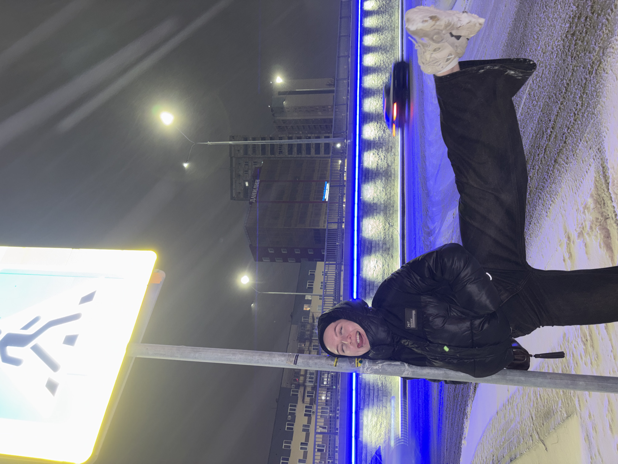 At night in Perm, Russia, a person lies on their side against a pedestrian crossing signpost. They wear a black hooded jacket, dark pants, and off-white sneakers. The sign itself is brightly illuminated. In the background, buildings are visible, including one labeled "Данилин." A bridge with bright blue lights runs horizontally across the scene. A vehicle with illuminated tail lights is also on the bridge. Snow is visible on the ground. Streetlights dot the environment.