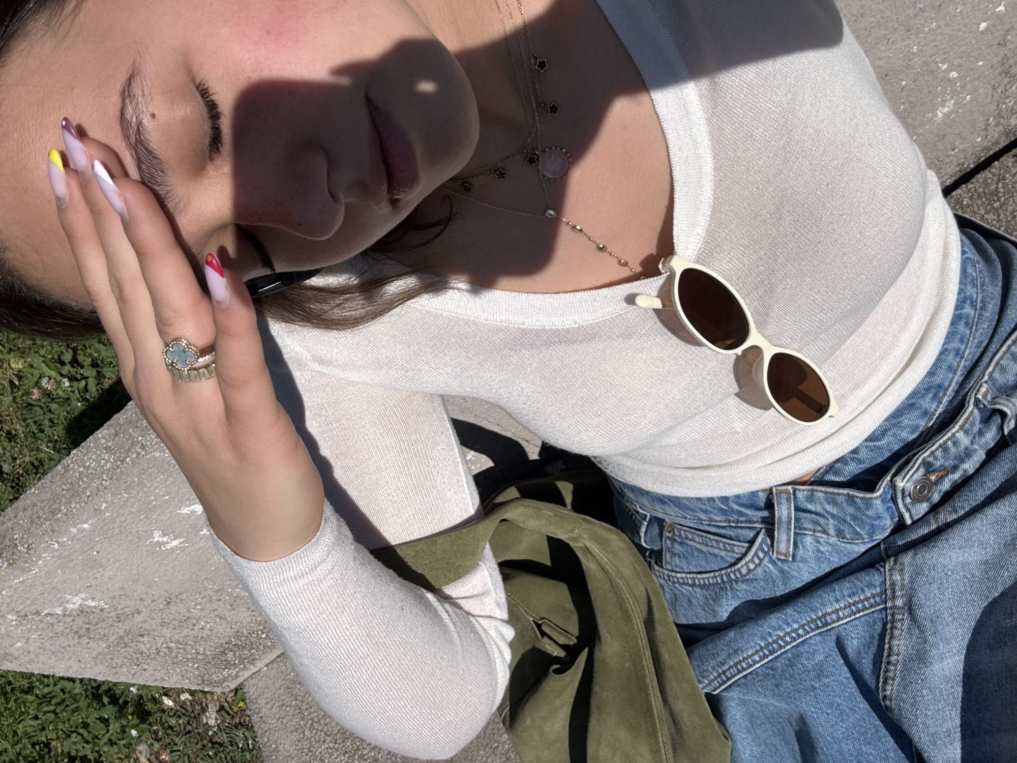 A woman rests in sunlight outdoors. She wears a white long-sleeved top, blue jeans, and multiple gold necklaces. A green suede jacket is draped near her lap. A ring adorns her hand, which is covering her face. Sunglasses rest on her shirt. Her nails are painted with a colorful design. The location appears to be in Milan, Italy, indicated by the style and materials of the concrete bench on which she is sitting, combined with the bright, Mediterranean sunlight. The setting is likely a park or garden within the city.