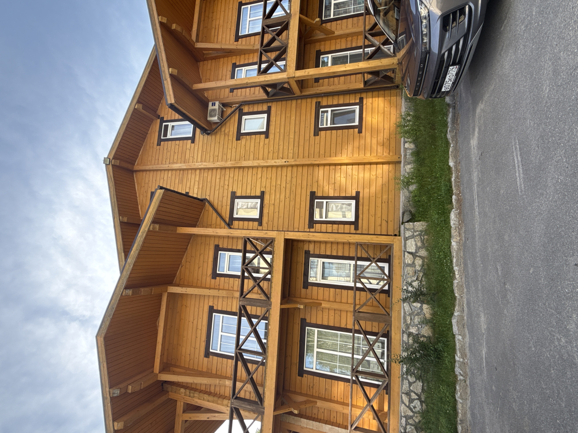 This is a vertically oriented, slightly angled shot of a wooden house in Syrostan, Russia, with a car parked nearby on an asphalt road. The house is multi-story, constructed with natural wood paneling and features numerous windows with dark frames. Decorative wooden beams and railings are visible, suggesting an architectural style that emphasizes natural materials. The windows on the upper floors are smaller and square, while those on the lower levels are larger and rectangular. An air conditioning unit is mounted on the exterior wall of the house.
In the foreground, to the right of the house, is a dark-colored car with a license plate that reads "B075KO 774". The car is parked on an asphalt surface, which transitions into a narrow strip of green grass and then a low wall made of natural stones. The lighting suggests it is daytime, with the sun casting soft shadows, possibly late afternoon or early morning. The sky is visible in the upper left of the frame and appears partly cloudy. The overall impression is one of a residential building in a natural setting, likely a rural or semi-rural area, given the prominent wooden architecture and surrounding greenery. No people are visible in the frame, and no specific activity or event is depicted, other than the presence of the parked car. The scene appears peaceful and quiet.