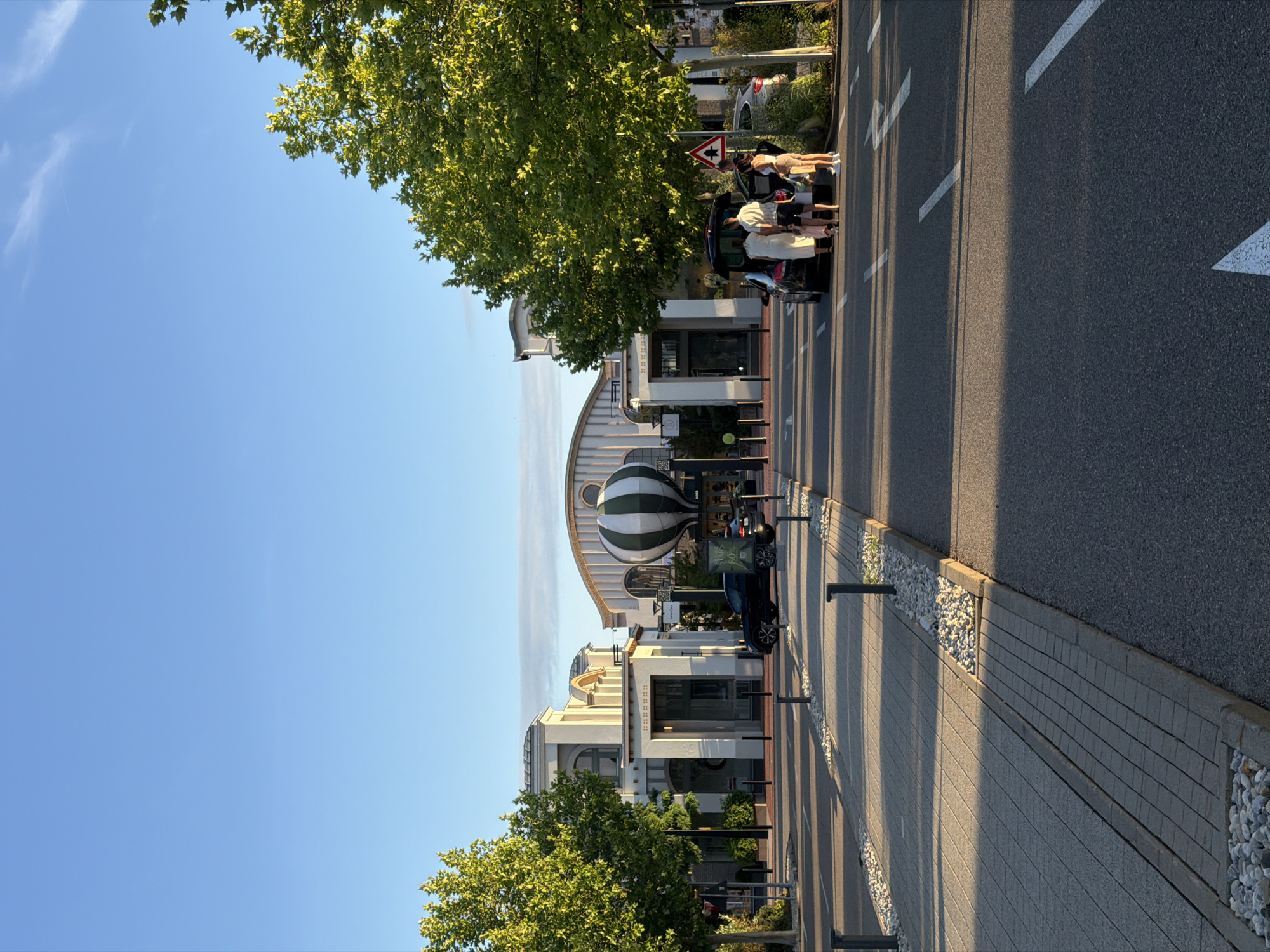 The image depicts a wide-angle outdoor view of a modern architectural complex featuring clean lines and light-colored facades, possibly a shopping outlet or a business park, situated in Ingolstadt, Germany. The sun casts long shadows across the paved pathway and asphalt road, indicating it is either early morning or late afternoon. A prominent, large hot air balloon, striped in green and white, is visible in front of one of the buildings.
To the right of the frame, a group of four individuals, two adults and two children, are gathered around the open trunk of a black SUV. One adult, a woman in a light-colored dress, appears to be loading items into the vehicle. Another adult, a man in shorts and a t-shirt, is standing nearby. The other two individuals, seemingly children, are also near the car. A second black car is parked to the right of the SUV, and a triangular road sign with a warning symbol is visible on a pole.
On the left side of the image, a tree with vibrant green foliage shades a portion of the pathway. The pathway itself is paved with interlocking bricks and bordered by a strip of small white pebbles and dark grey curbs. Several black bollards are spaced along the edge of the pathway.
The sky is clear and blue, with a few wispy clouds visible in the upper portion of the image. The overall scene suggests a pleasant day in a well-maintained urban or suburban environment. The specific location is identified as the Ingolstadt Village, a luxury outlet shopping center.