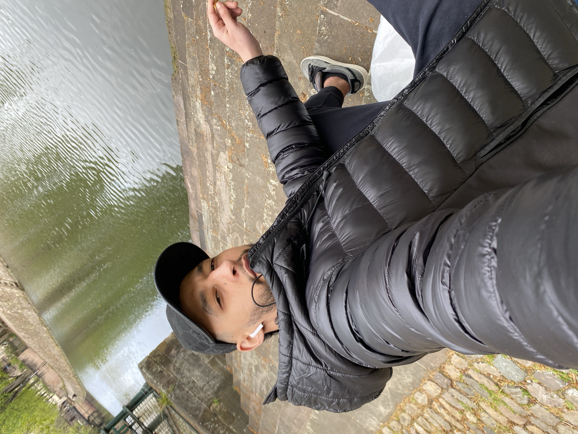 A man stands by a canal in Strasbourg, France, holding an object in his right hand. He wears a black puffer jacket, black cap, and grey sneakers. In his ear, he has an earbud. The canal has green water with ripples, and its edge is marked by a stone barrier. The cobblestone ground is visible around the man.