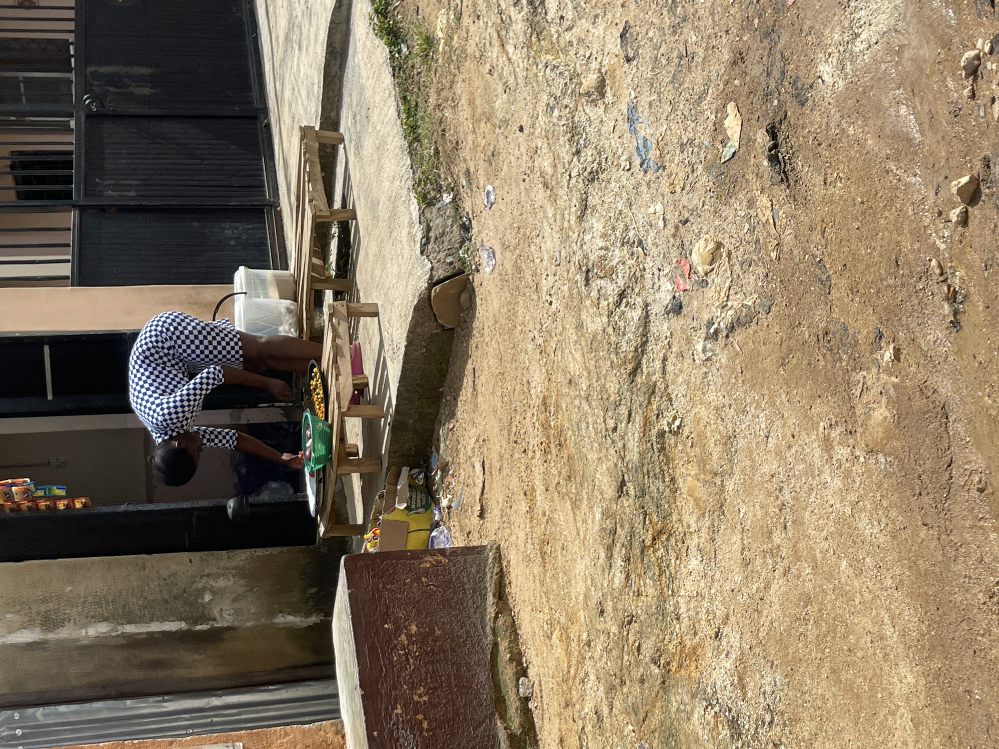 A young woman wearing a blue and white checkered dress bends over a low wooden table, preparing food. She is standing in an outdoor environment with a dirt and gravel ground, littered with some plastic bags and debris. Behind her, a black metal gate is visible, along with a building with orange and grey walls. The scene appears to be in a marketplace or an informal commercial area in Jos, Nigeria, judging by the overall setting and the presence of a food stall setup.
