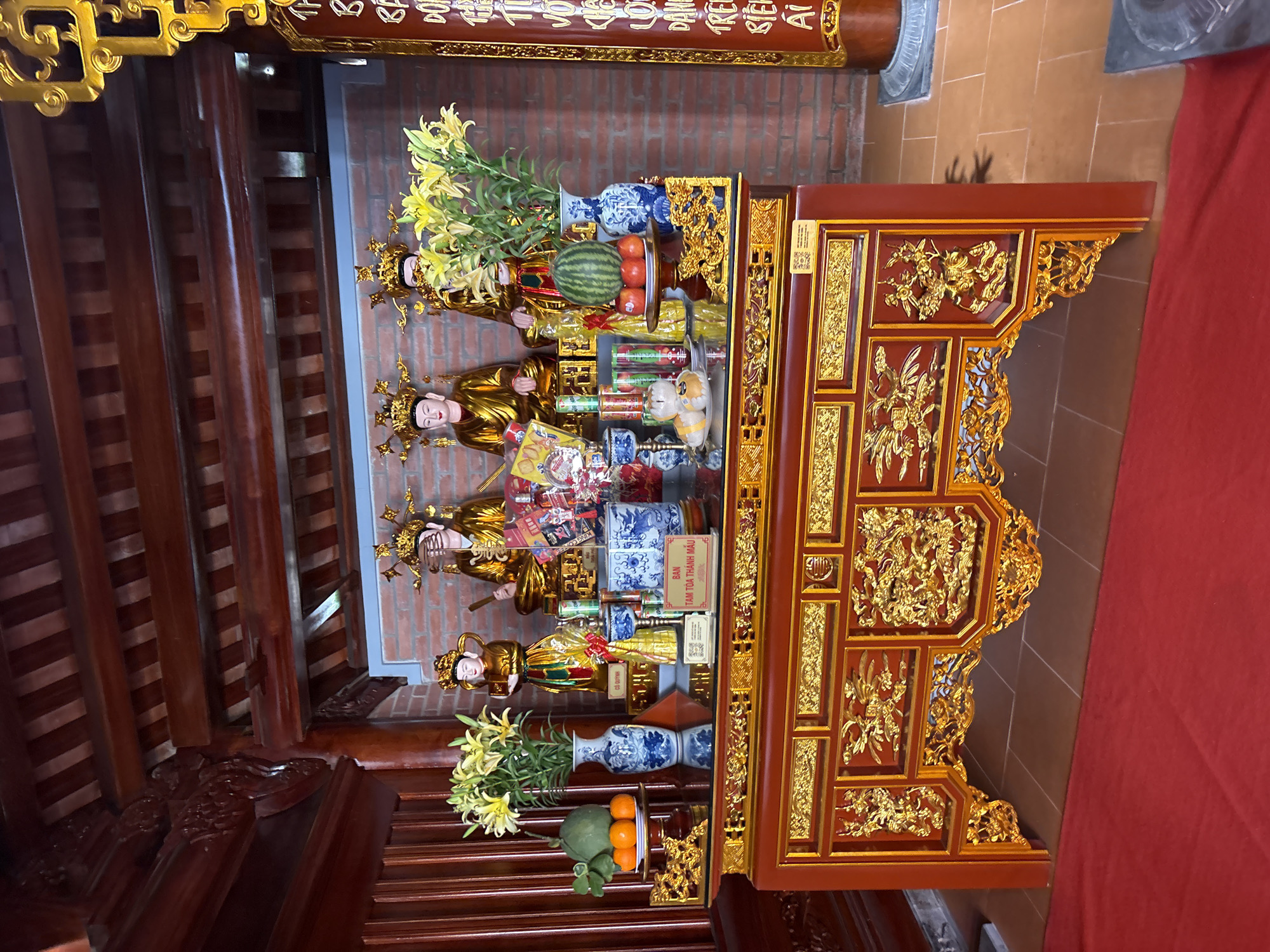 The image depicts an altar inside a building, likely a temple or shrine, located in Diên Khánh, Vietnam. The altar is elaborately decorated in red and gold, with intricate carvings. On the altar, there are offerings, including fruit, flowers, and small statues of deities. These statues are adorned with golden crowns and robes. Ceramic vases with blue and white patterns are also present. The background reveals a brick wall and wooden beams of the building's structure. A red cloth hangs near the altar.