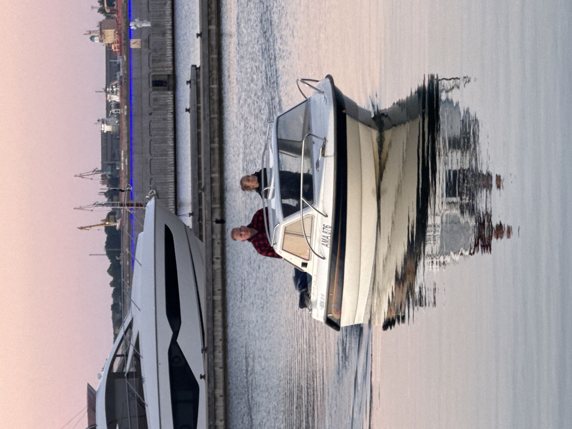 The image depicts a marina in Tallinn, Estonia, during a twilight hour, as indicated by the soft pink hues of the sky. In the foreground, a small white motorboat with the registration "AMA 876" is navigating the calm water, leaving a gentle wake. Two individuals are onboard: a man with short blond hair, wearing a red and black checkered shirt, is seated at the helm. A woman with blonde hair tied back, wearing a dark top, stands beside him, looking towards the left of the frame. Behind them, a larger white yacht with dark accents is moored. The background features a harbor wall with industrial cranes visible in the distance. The water reflects the boats and the sky, creating a serene and slightly diffused image.