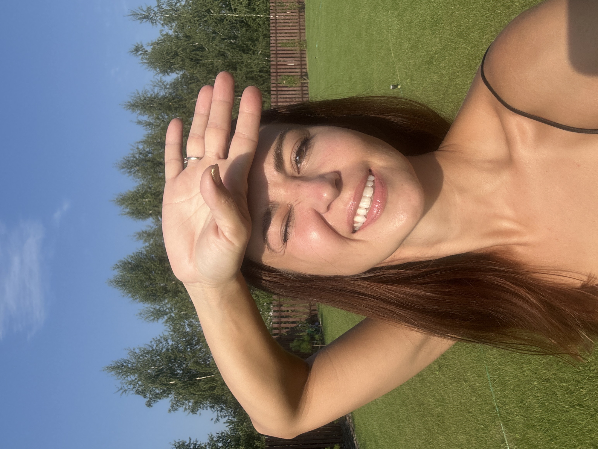 A woman with brown hair is taking a selfie outdoors. She is holding her right hand up to her forehead, shielding her eyes from the sun. She is smiling and winking. The background is a green lawn with a wooden fence and trees. The sky is blue with some clouds. The location is identified as Samarsky, Russia.