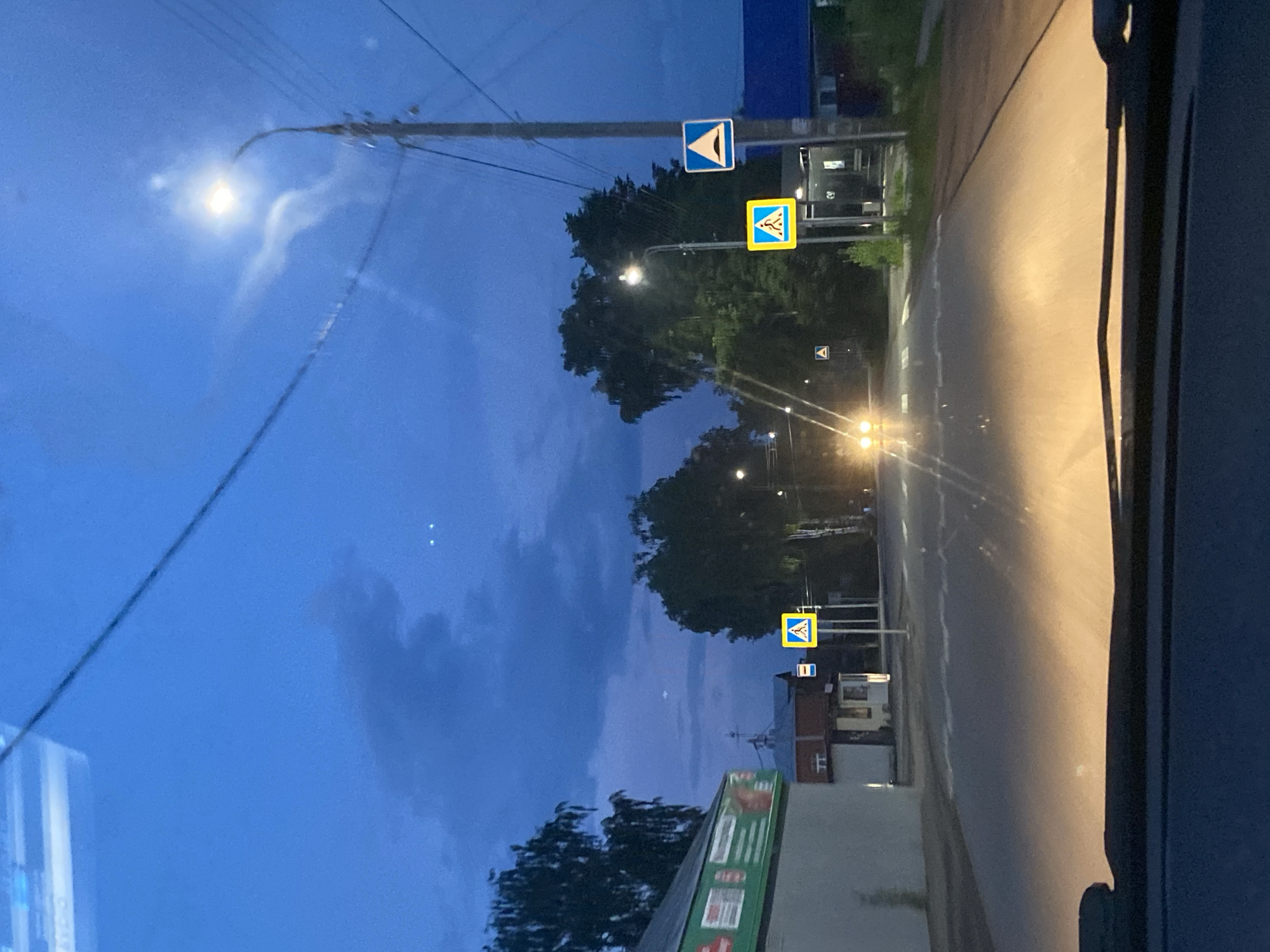 The image depicts a road scene at dusk or night, likely in Votkinsk, Russia. In the foreground, the dashboard and windshield of a vehicle are visible, with headlights illuminating the road ahead. Several road signs are present, including two pedestrian crossing signs and a speed bump warning sign. The road itself appears to be asphalt with visible lane markings.
On the left side of the road, a building with signage, partially obscured, is visible. Further down the road, trees and other buildings can be seen. The sky is dark blue with clouds and a bright street light illuminates the scene. Two distant, small lights are also visible in the sky, possibly aircraft or stars. The overall atmosphere suggests a quiet, rural or suburban setting during twilight. There are no people or animals clearly visible in the image.