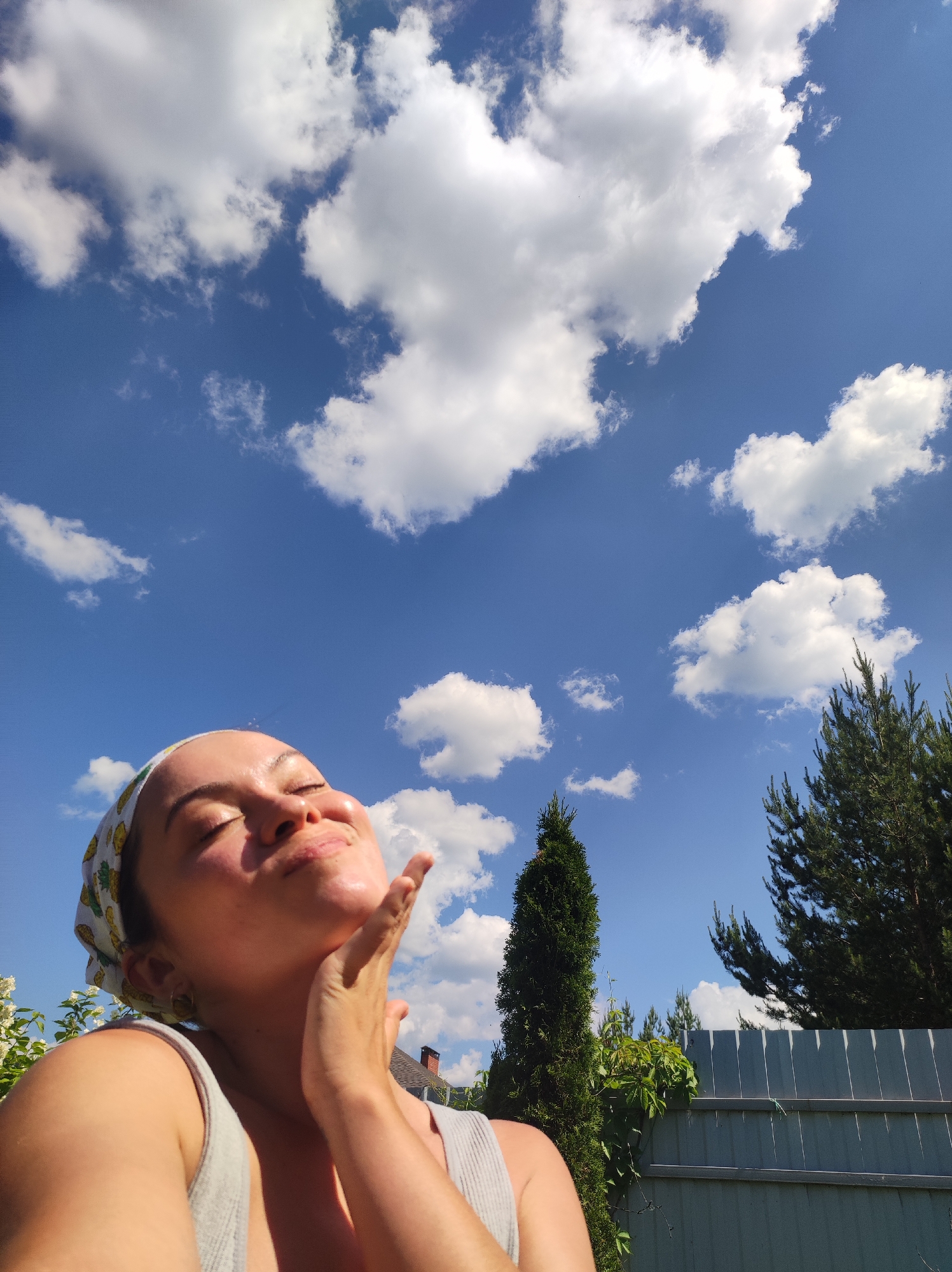 A woman is pictured outdoors, her face tilted upwards towards a bright blue sky filled with white, puffy clouds. She is wearing a bandana with a pineapple pattern and a grey tank top. Her eyes are closed, and her hand is gently touching her chin. The background shows greenery, a metal fence, and trees, suggesting a suburban or rural setting. The presence of a house's roof is visible behind the woman, and evergreen trees are prominent in the mid-ground and background. The lighting indicates it is daytime, likely sunny. The location is specified as Фоки, Russia.