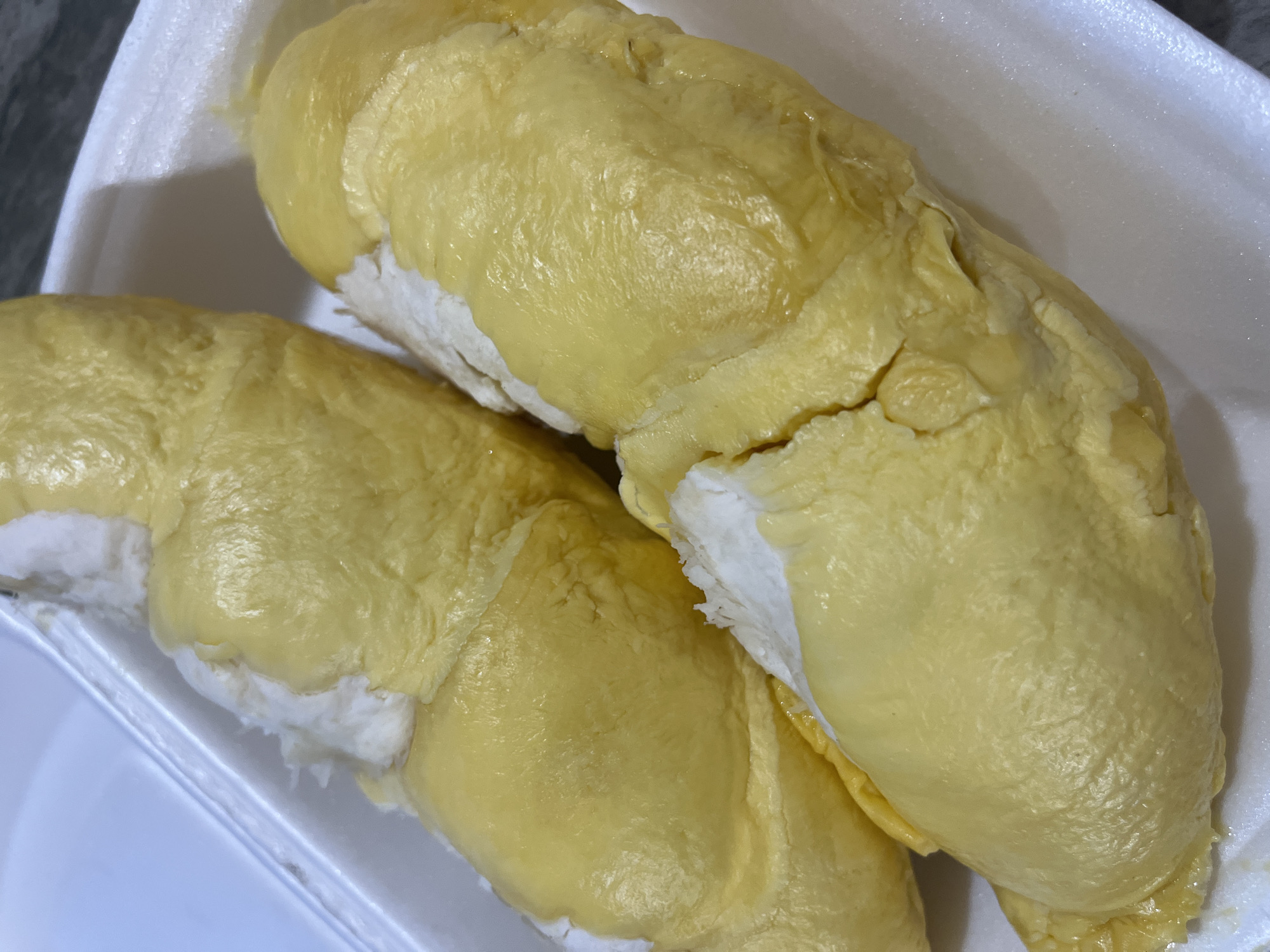 The image shows two segments of durian fruit in a white styrofoam container. The durian flesh is a pale yellow color and appears soft and creamy. The container is sitting on a surface, partially visible at the bottom left. The background is a dark gray color, possibly a countertop or wall. The image was taken in Bac Nha Trang Ward, Vietnam.