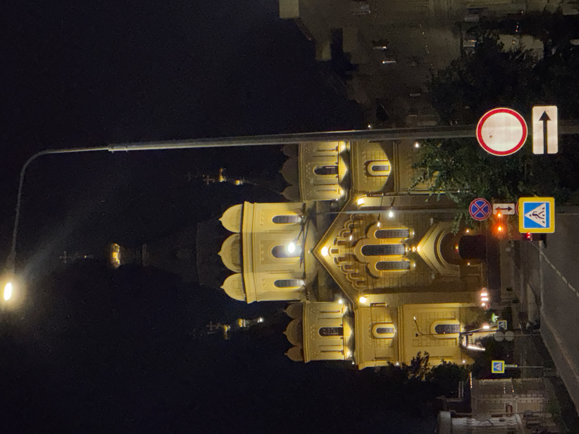 At night in Nizhny Novgorod, Russia, an illuminated yellow church with multiple domes and a tall steeple stands prominently. A street lamp with a bright bulb extends over the scene, casting light onto the dark sky and the building. The church features arched windows and decorative architectural elements, all aglow with warm light. In the foreground, a street is visible, with several traffic signs. These include a pedestrian crossing sign, a circular sign with a red border and a horizontal white bar (indicating no entry), and another sign showing a right arrow. The surrounding area is dark, with the indistinct shapes of other buildings and trees visible in the background. The overall impression is one of a well-lit landmark on a quiet city street at night.
