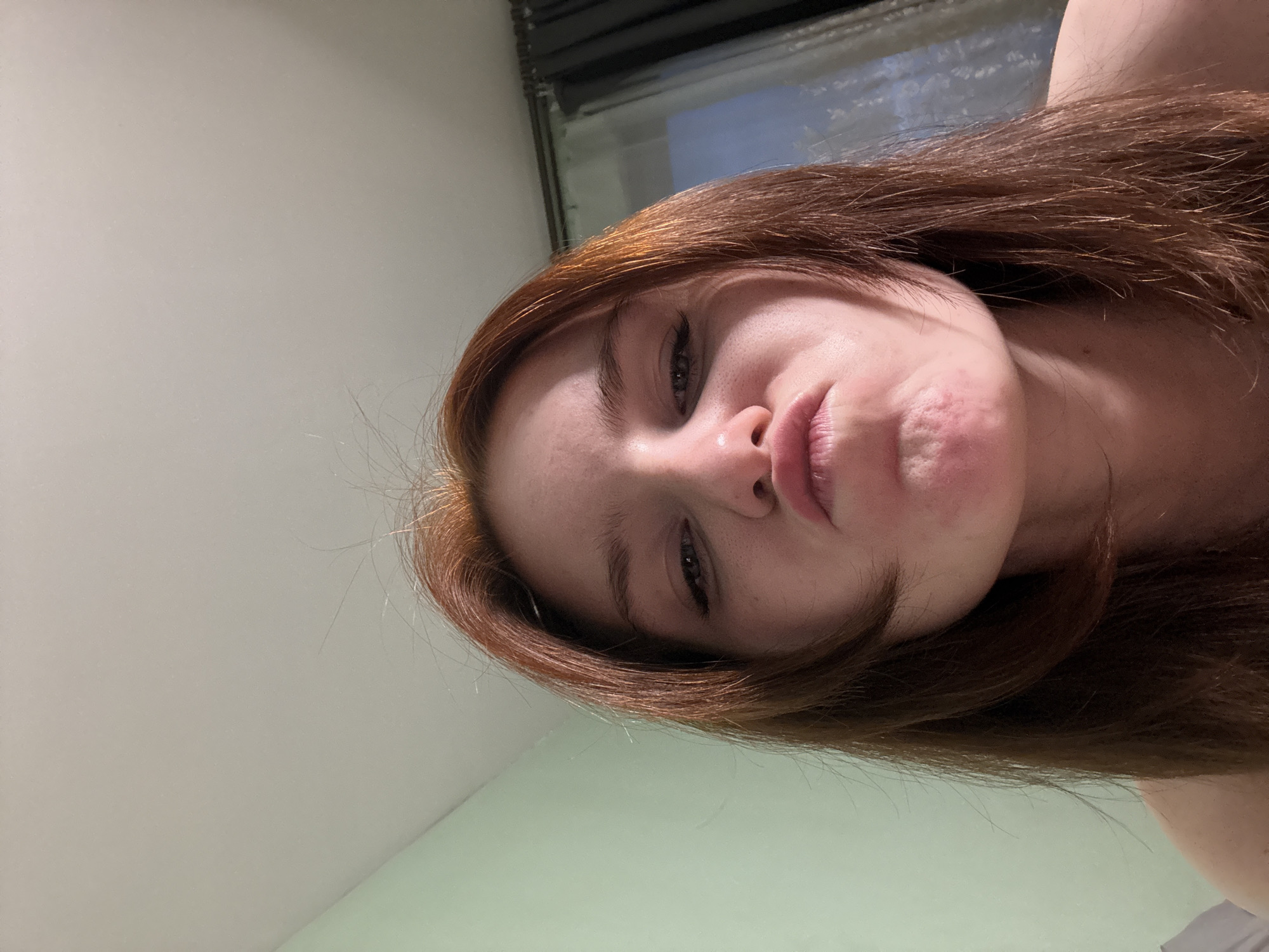 A close-up, high-angle shot focuses on a fair-skinned woman with red-brown hair and a slightly puckered mouth. The background is divided into two sections: a white wall on the left and a muted green wall at the bottom. At the top is a window. The location is Unknown, Unknown.