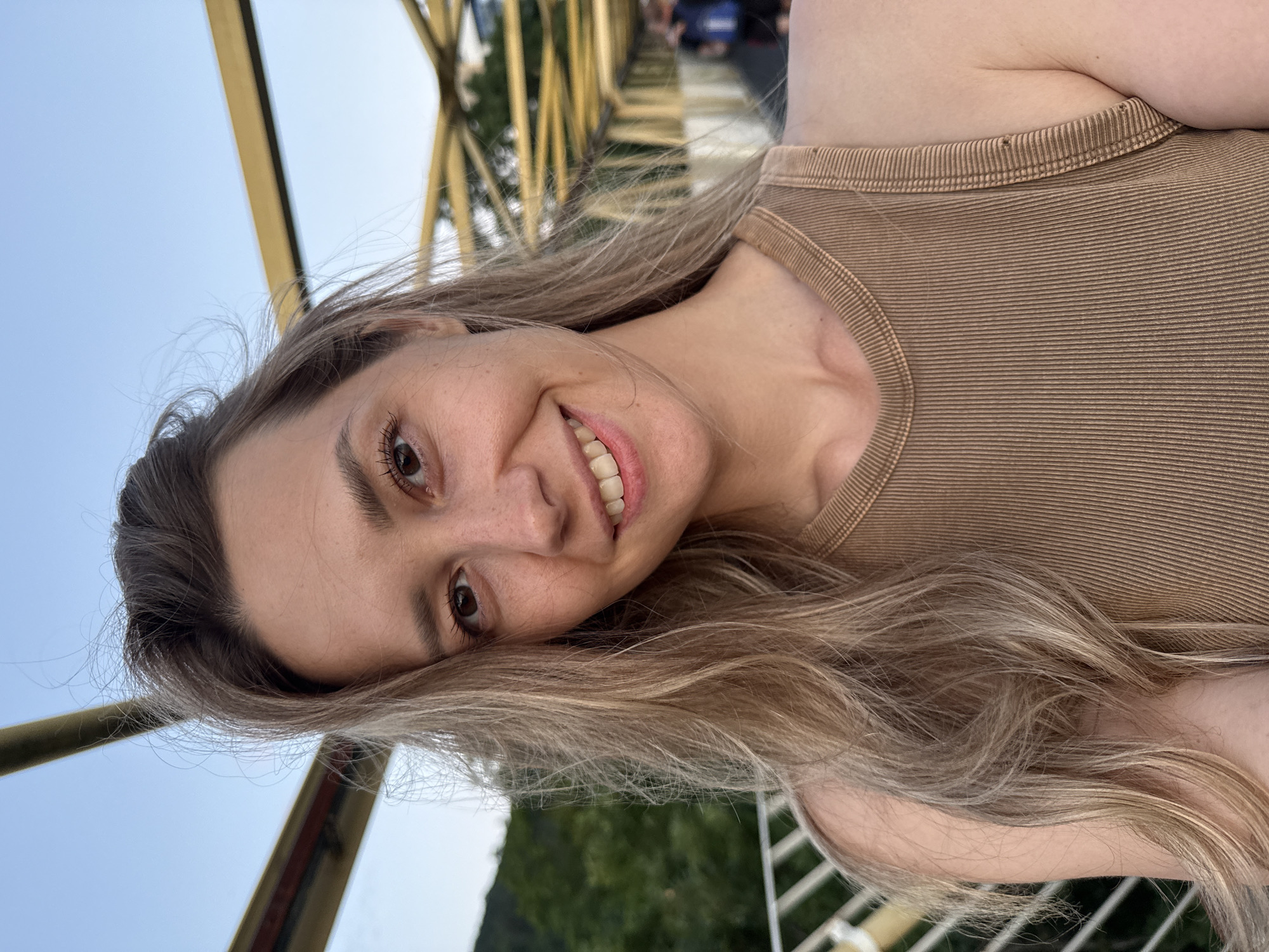 A woman with long blonde hair and freckles is taking a selfie on a bridge with a yellow railing. She is wearing a brown ribbed tank top and smiling at the camera. In the background, trees and a body of water can be seen. The bridge appears to be in an urban area, possibly in Nebug, Russia, as indicated by the context.