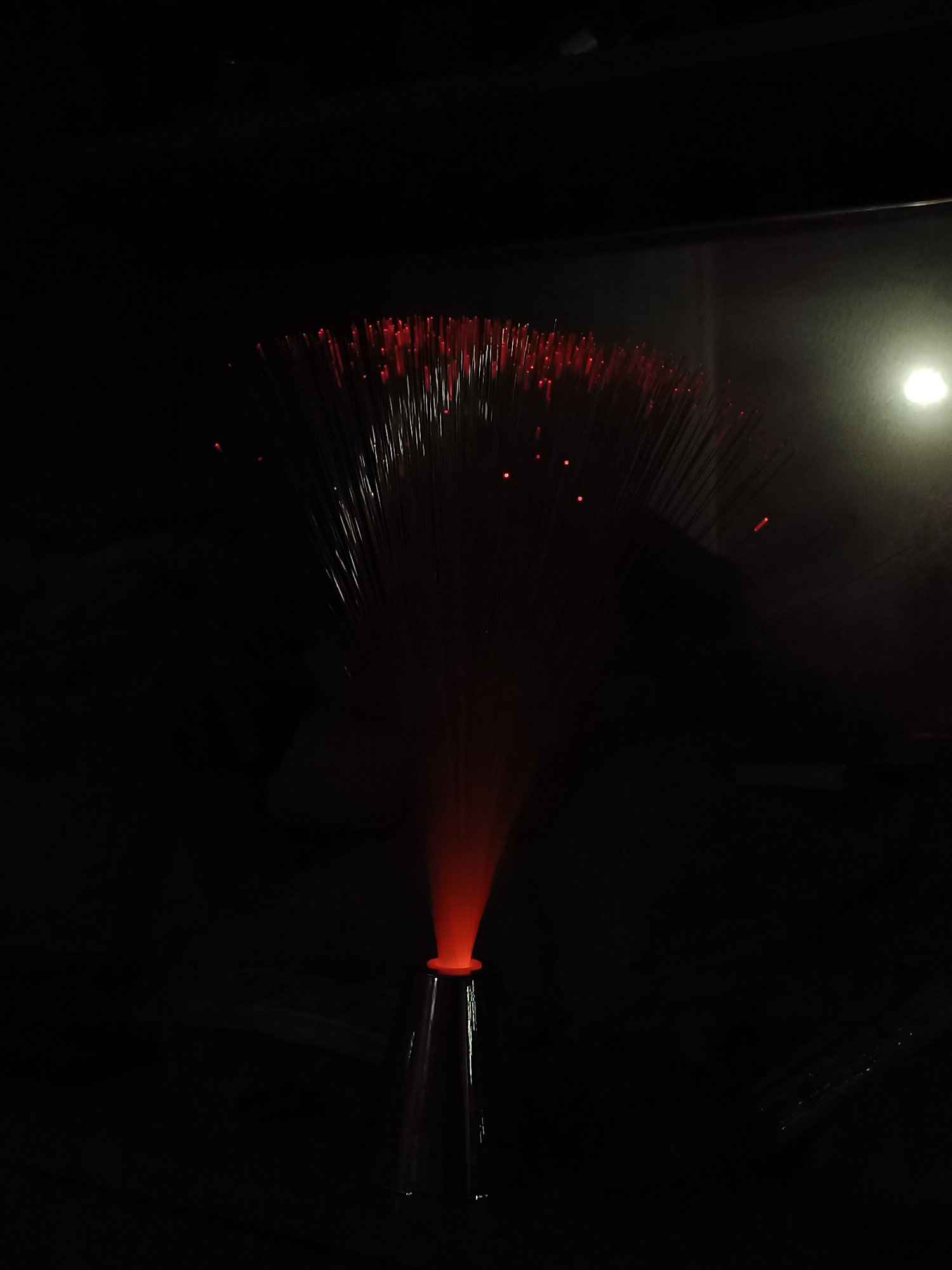 A dimly lit photograph shows a fiber optic lamp. The lamp has a base that flares out at the top. The fibers extend upwards and outward, with small points of red light visible at the tips of many fibers. The background is dark and indistinct, but a faint light source is visible in the upper right. The location in Cherkovitsa, Bulgaria is not discernible from the image.