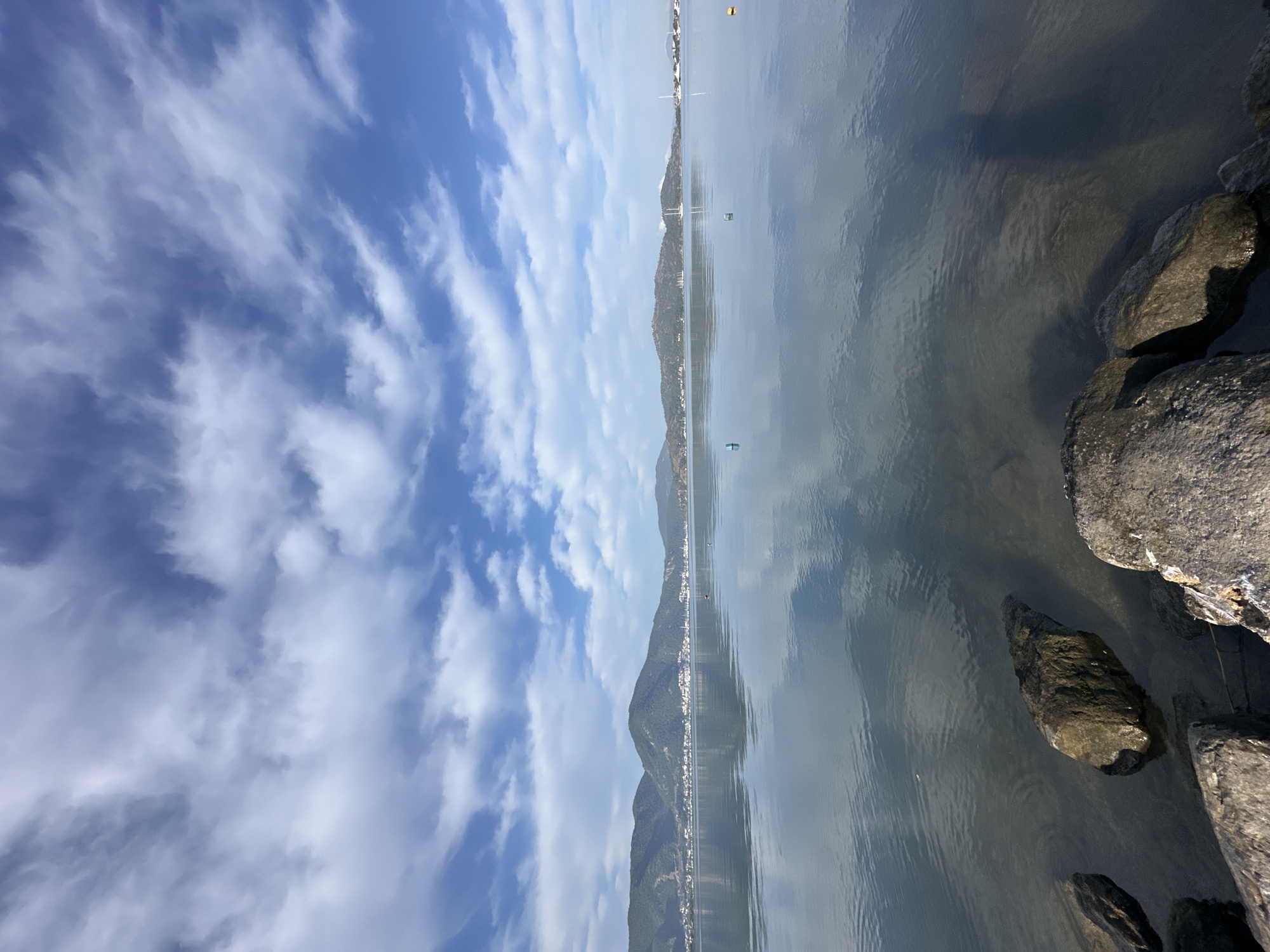 The image depicts a waterfront scene in Fethiye, Turkey. A body of water, possibly the Mediterranean Sea or a bay, occupies the majority of the right side of the frame, with reflections of the sky and distant mountains visible on its surface. Rocks line the edge of the water on the right. Across the water, a town stretches along the base of a mountain range. The left side of the image is dominated by a blue sky with streaks of white clouds. The scene appears to be captured during daylight hours. There is no obvious evidence of people in the image.