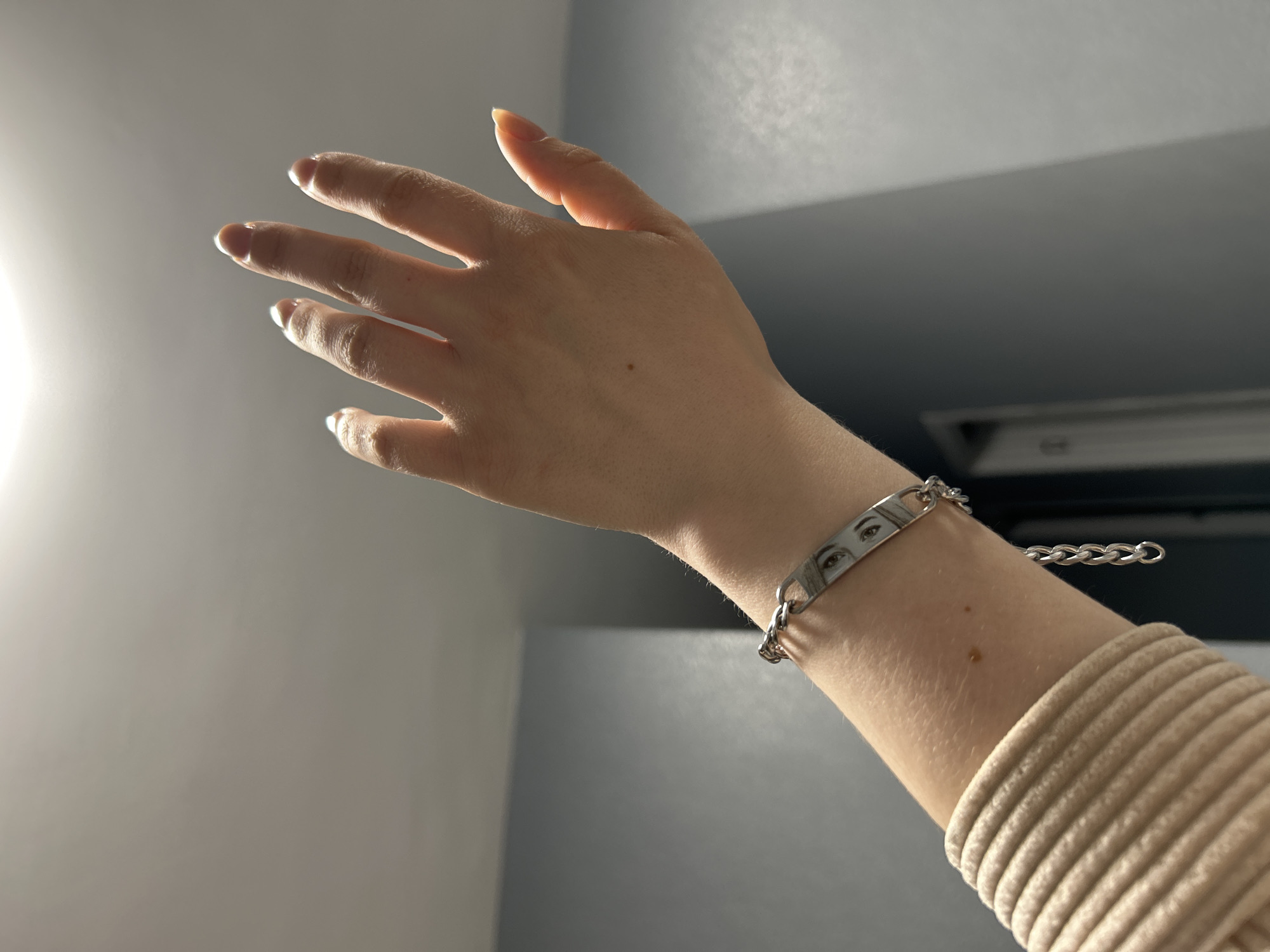 A person's arm and hand are extended upward against a blurred gray background. The hand is open with fingers slightly spread, and the fingernails appear to be painted white. A silver-colored bracelet is on the wrist, featuring a rectangular centerpiece with two human eyes imprinted on it. The bracelet has a chain attachment. The arm is wearing a tan or light brown ribbed garment. There are no specific indicators to pinpoint the location within Saint Petersburg, Russia. The only light source is a glowing orb on the left of the image.