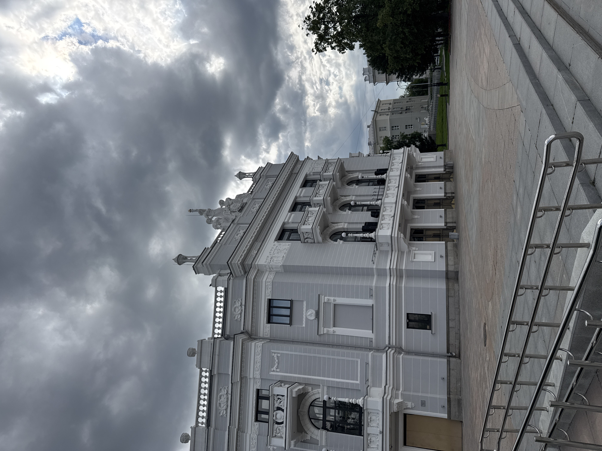 The image shows the facade of a large, ornate, light gray building with white decorative elements, likely a public building or theater, under a dramatic, cloudy sky. The building features multiple balconies with intricate railings, arched windows, and sculptural details along the roofline. In front of the building is a paved plaza with a set of wide stone steps leading up to a ramp with polished metal railings. To the right of the building, several trees are visible, and in the background, other multi-story buildings can be seen. The overall scene suggests an urban environment in Yekaterinburg, Russia, possibly the Yekaterinburg Opera and Ballet Theatre, given its architectural style and prominent location. There are no people visible in the image.