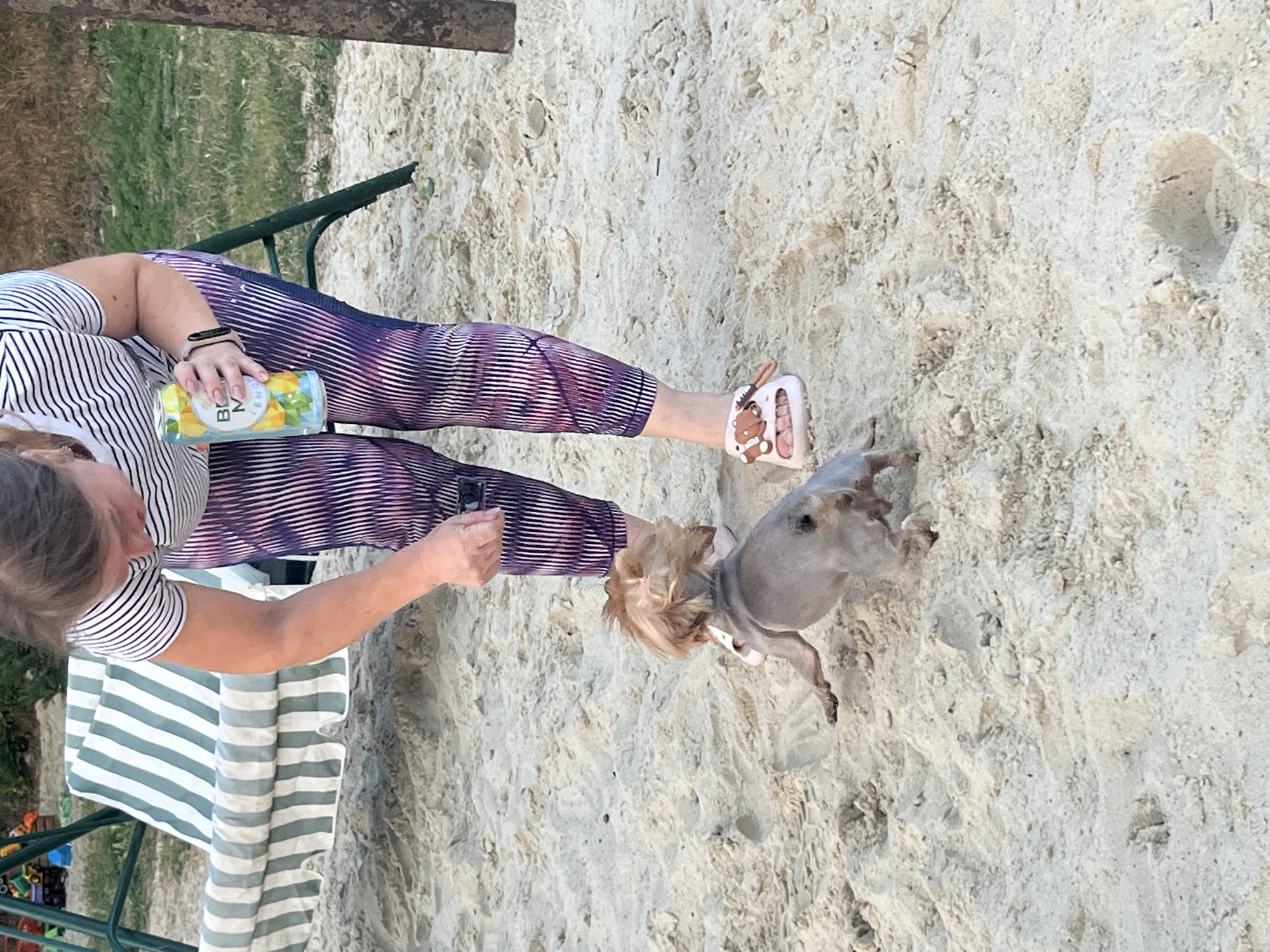 A woman is on a sandy area, bent over, holding a can of "B.M. Lemon" in her right hand. She is wearing a striped t-shirt and patterned leggings. In front of her, a small dog with light brown fur is facing away from the camera, appearing to be moving or playing on the sand. A swing set with a striped cushion is visible in the background to the left, along with some children's toys. To the right, a tree trunk is partially visible. The ground is covered in sand and some scattered green vegetation is present. The location is the city of Lozuvatka, Ukraine.