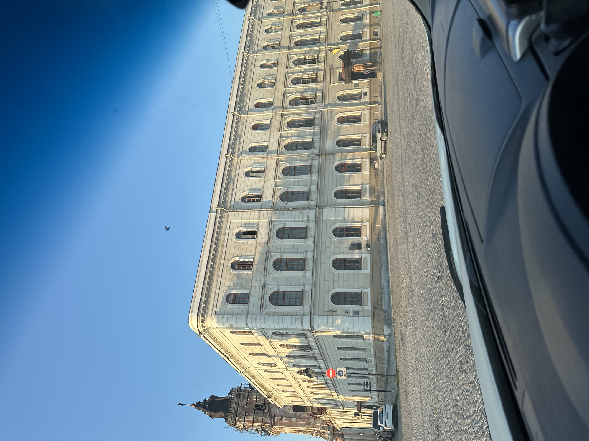 The image captures a view of the Trinity Cathedral in Kamianets-Podilskyi, Ukraine, from inside a vehicle. The sun casts long shadows across the cobblestone square in front of the cathedral, indicating it is either early morning or late afternoon. The grand, pale yellow facade of the cathedral, with its numerous arched windows, dominates the right side of the frame. To the left, the tower of the cathedral, partially covered in scaffolding, reaches towards the clear blue sky. A single bird is visible flying high above the buildings. Several cars are parked on the square, and a traffic sign with red and white markings is situated near the cathedral entrance. The foreground shows the dashboard and windshield of a car, suggesting the photograph was taken by a driver or passenger.