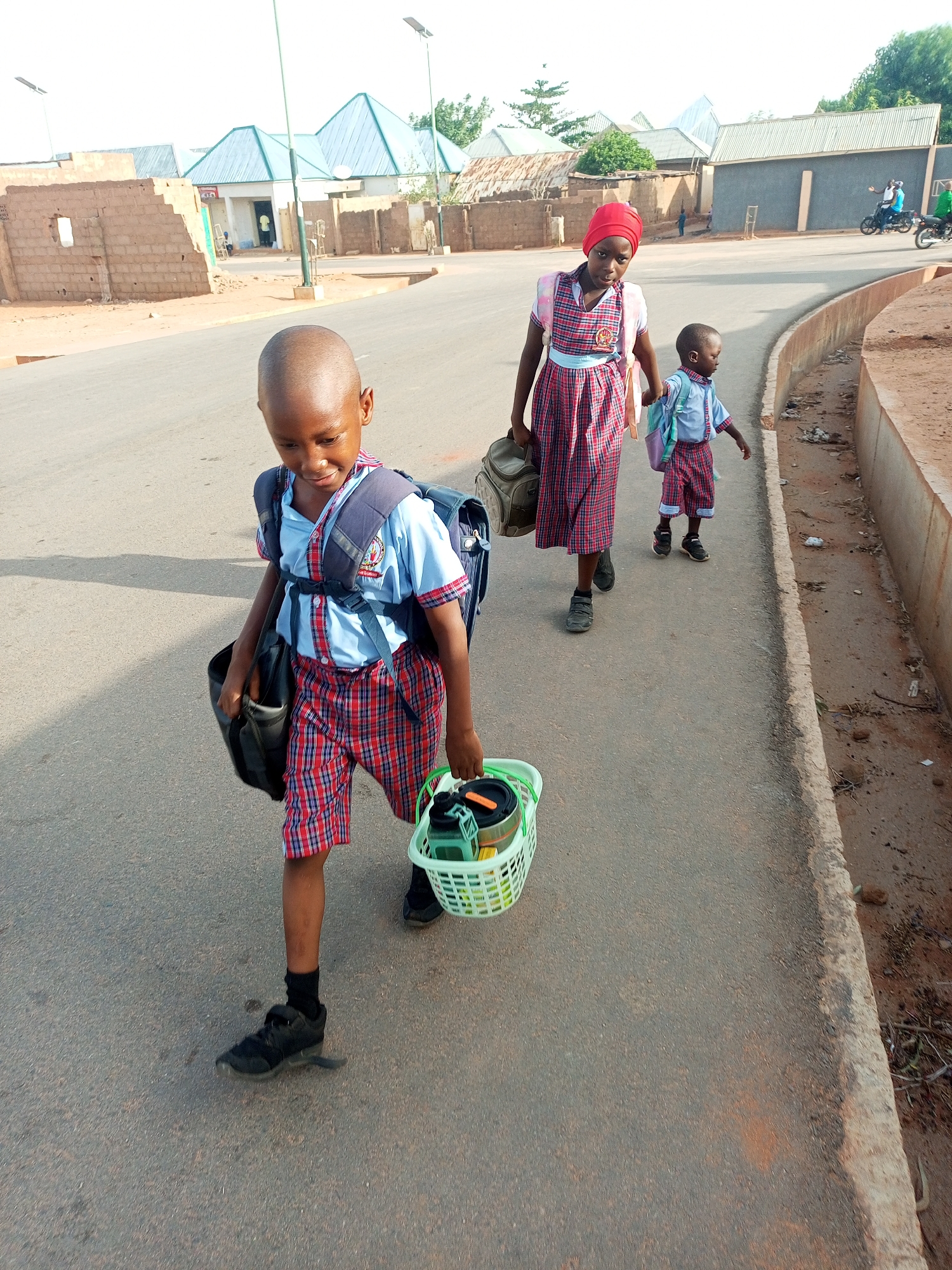 Three children in school uniforms walk on a paved road in Tempure, Nigeria. The child in the front carries a backpack and a basket filled with items. The child behind him is a girl wearing a red headscarf and carrying a bag. She is holding hands with a younger child who also has a backpack. The background includes buildings and a motorcycle on the street.