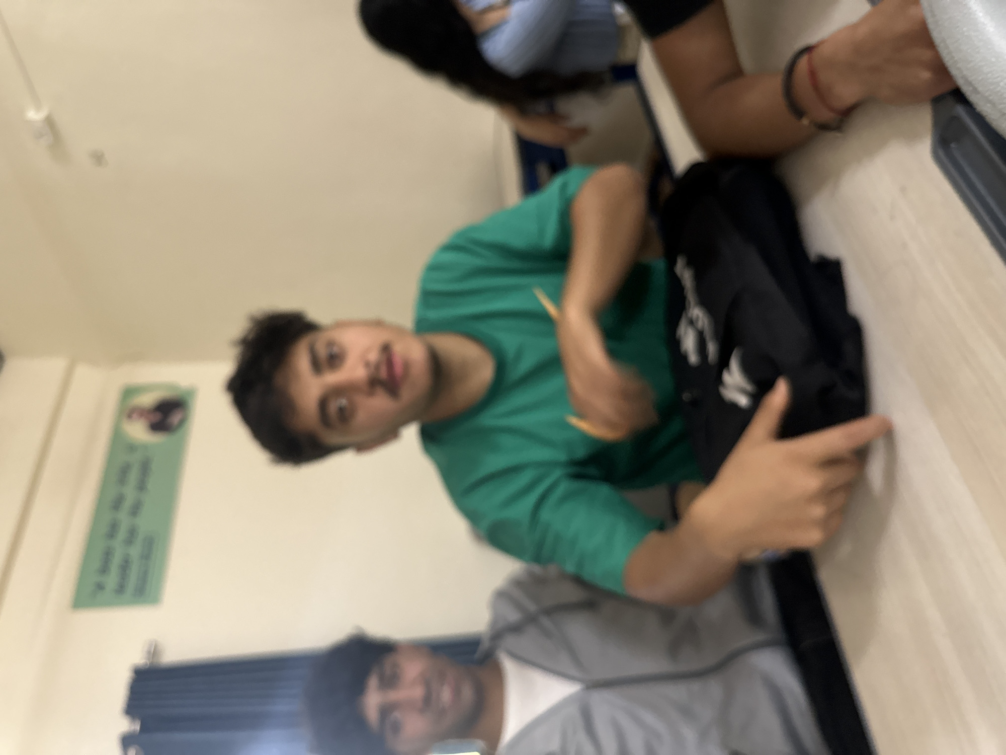 The image depicts a group of young people sitting at a desk in what appears to be a classroom or common area. In the foreground, two males are visible. The male on the left wears a grey hooded jacket over a white t-shirt and has dark, curly hair. The male in the center, wearing a green t-shirt, is the primary focus. He has dark hair and a mustache, and is holding a yellow pencil in his right hand, which is resting on a black folded garment on the table. His left hand is also on the table, pointing towards the garment. To the right of the central figure, a female with long dark hair, wearing a light blue top, is partially visible. The background includes a wall with a motivational poster featuring a quote and a picture of a person. A blue curtain is visible on the left side of the image. The environment suggests an educational or informal gathering space.