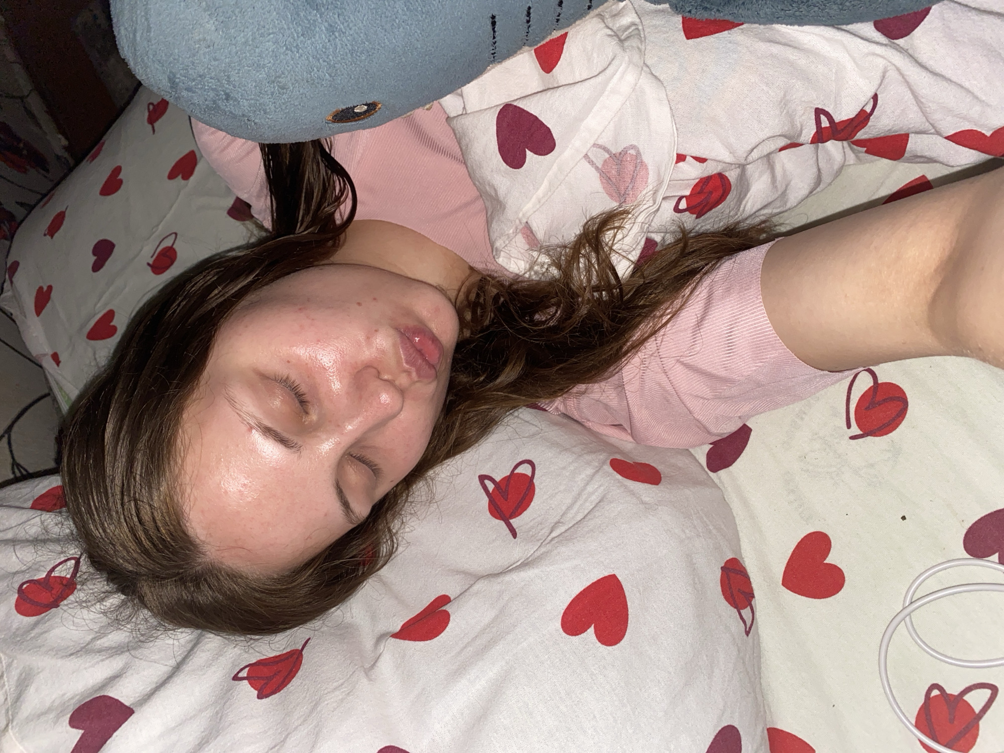 A young woman is lying in bed, asleep, with her head on a pillow patterned with red and maroon hearts. She is wearing a pink shirt. A blue stuffed animal is next to her. Her facial expression suggests she is sleeping deeply, with her lips pursed. The room appears to be a bedroom, with bedding and a wall visible in the background. The location is not specified in the image or context.