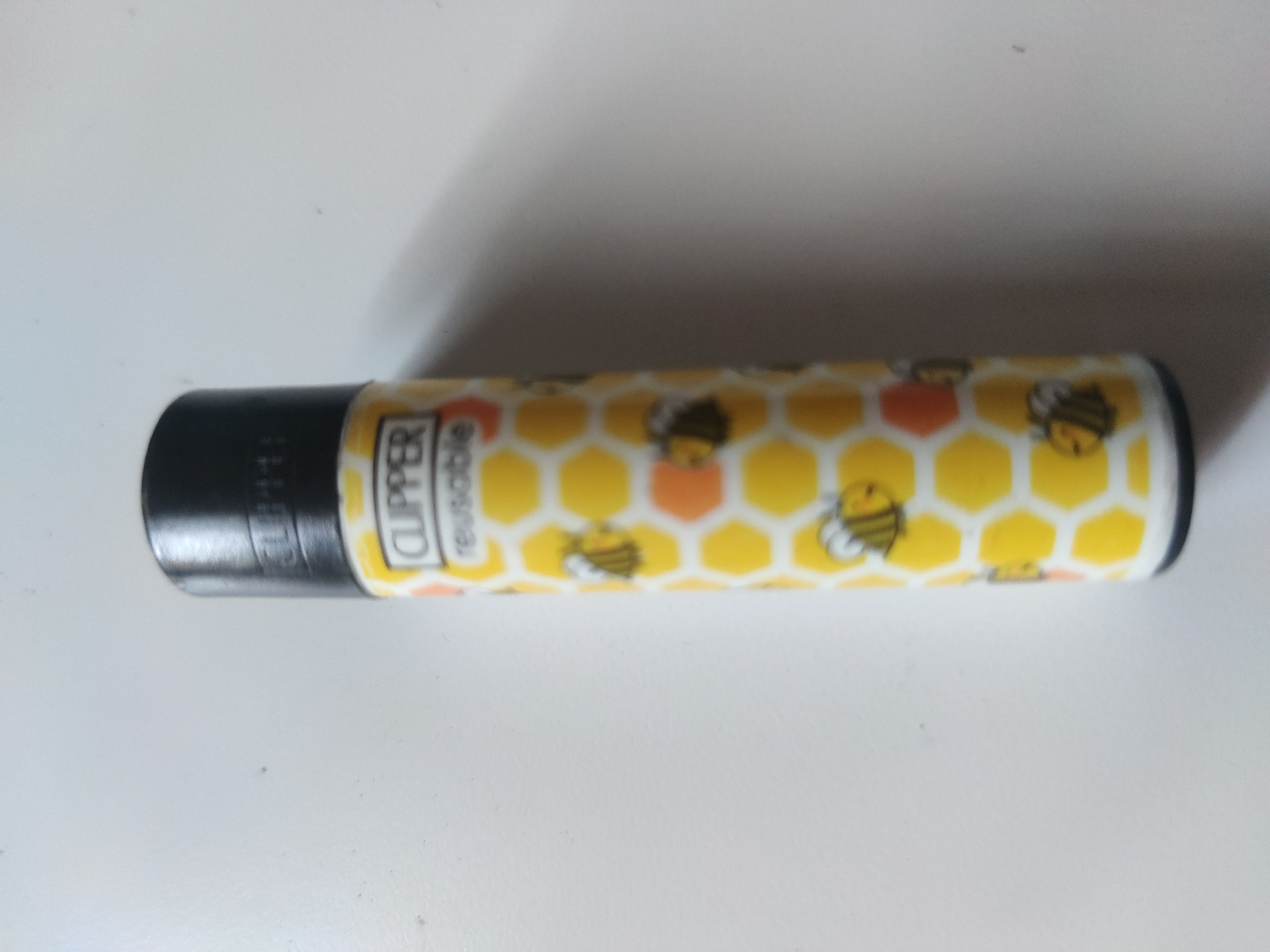 A cylindrical Clipper lighter with a black top is laying horizontally on a white surface. The body of the lighter is decorated with a pattern of yellow honeycombs and cartoon bees. The lighter has the word "Clipper" and "reusable" printed on its side.