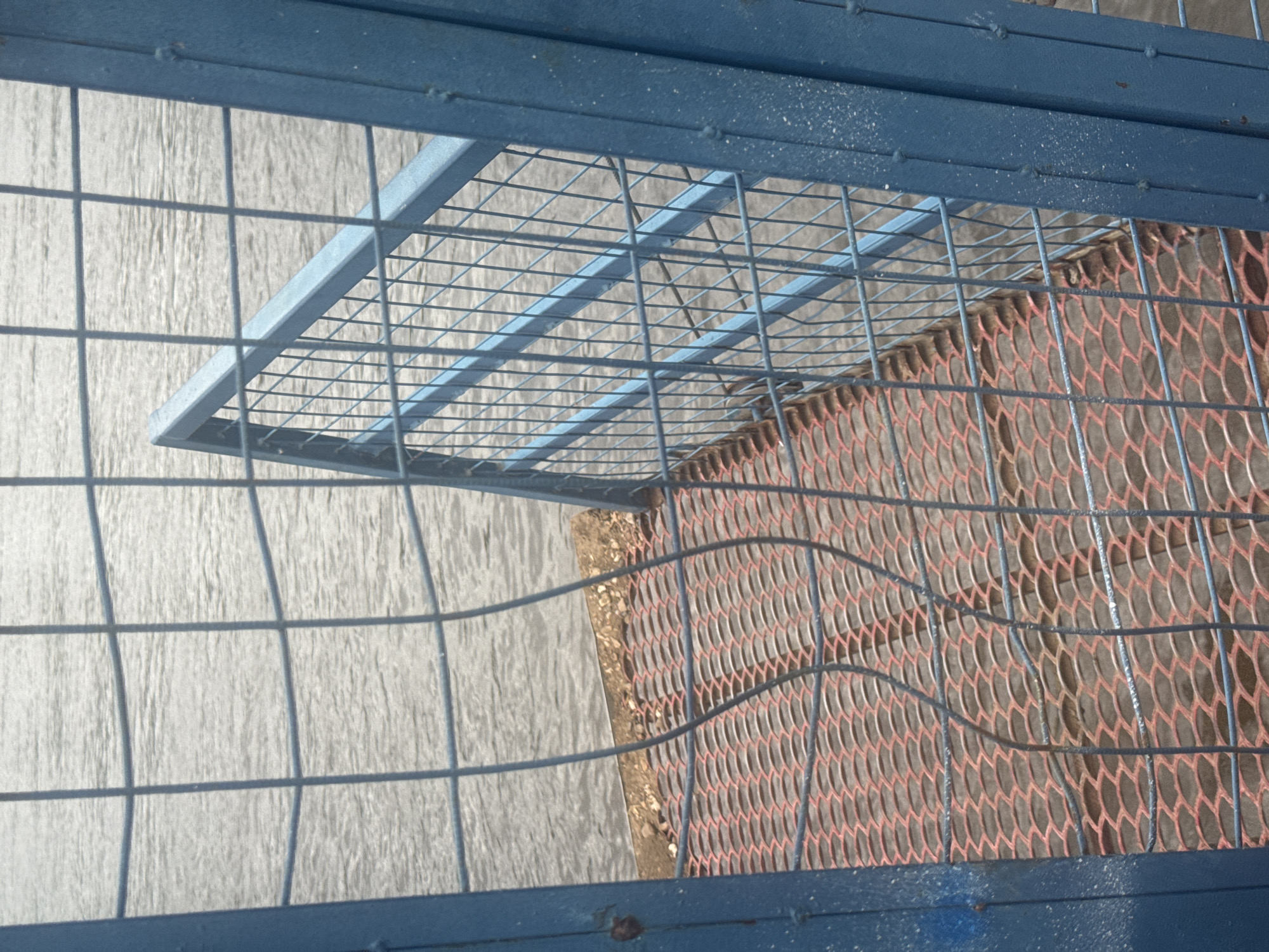 The image depicts a view through a blue metal fence of a dock or pier. The foreground shows a red expanded metal grating, common for walkways, leading towards the water. Beyond the grating, a concrete edge is visible, separating the walkway from the choppy gray water. A portion of a blue metal railing or barrier angles towards the water on the right side of the frame. The water appears calm with small ripples. The scene is set against a neutral, overcast sky. The location is identified as Ufa, Russia. There are no people or animals present. The overall impression is one of an industrial or public waterfront area.