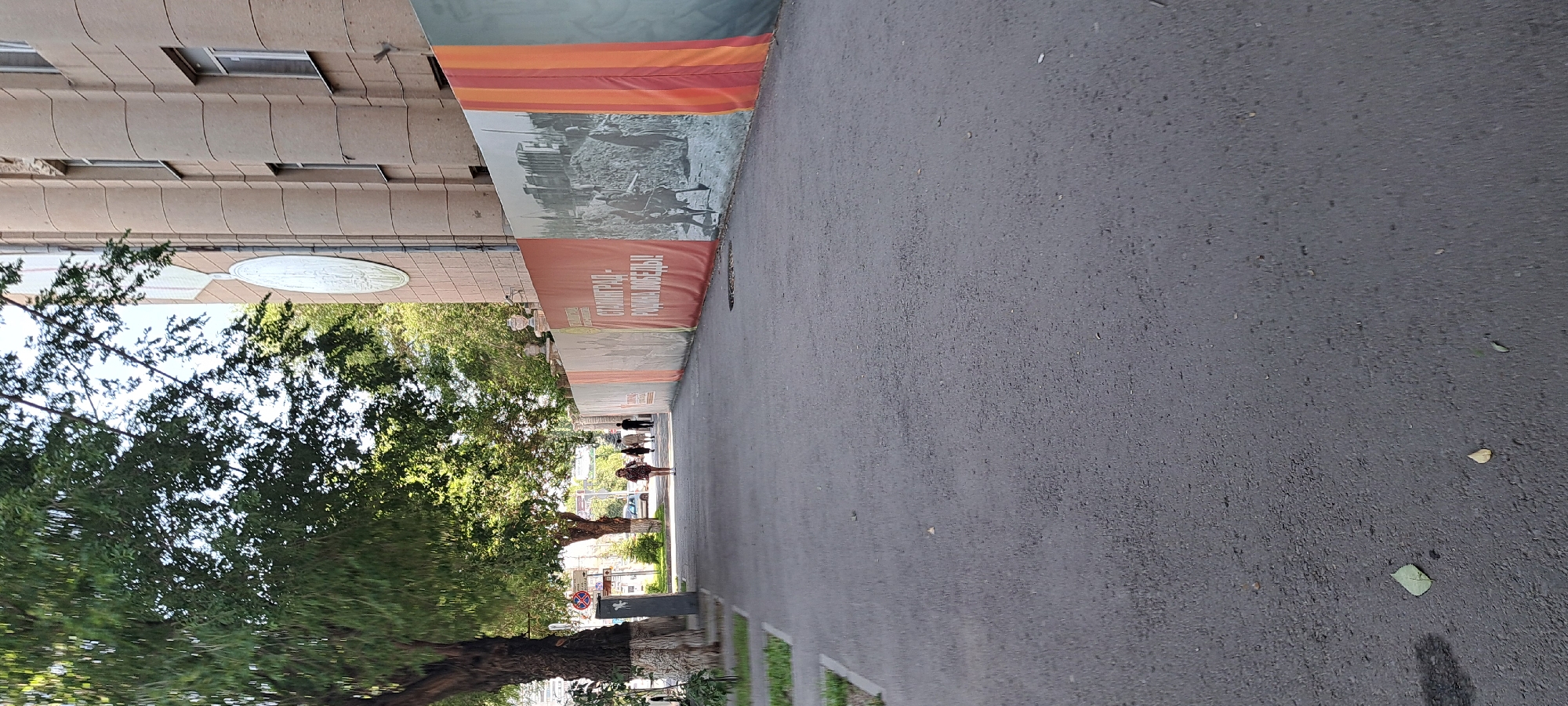 An eye-level, outdoor shot shows a wide, asphalt sidewalk on the left with a tree-lined path beyond it. A building with a beige facade and vertical columns is visible on the right. A large banner with a historical image and text runs along the building's base. Several people are walking away from the camera on the path. The sky is bright and overcast.