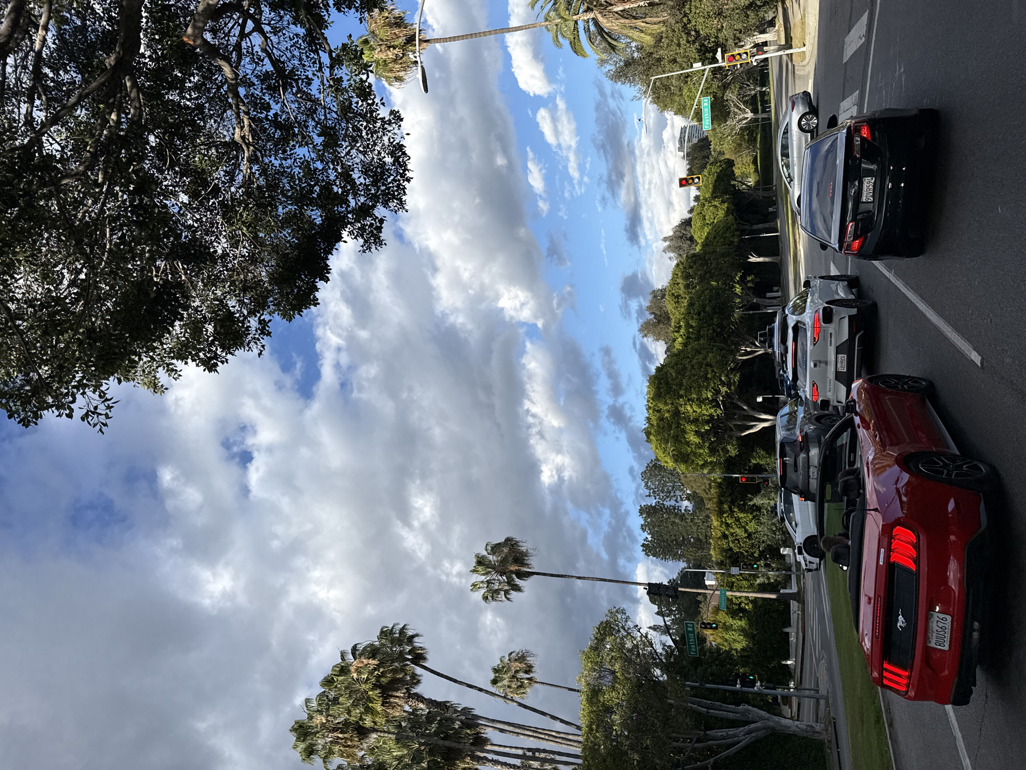 The image depicts a street scene in Beverly Hills, California, United States of America. Cars are stopped at a traffic light on Foothill Road. The sky is partly cloudy, with palm trees visible along the road. A red convertible Mustang is in the foreground, followed by several other vehicles including a black sedan. Street signs indicate the location as Foothill Road.