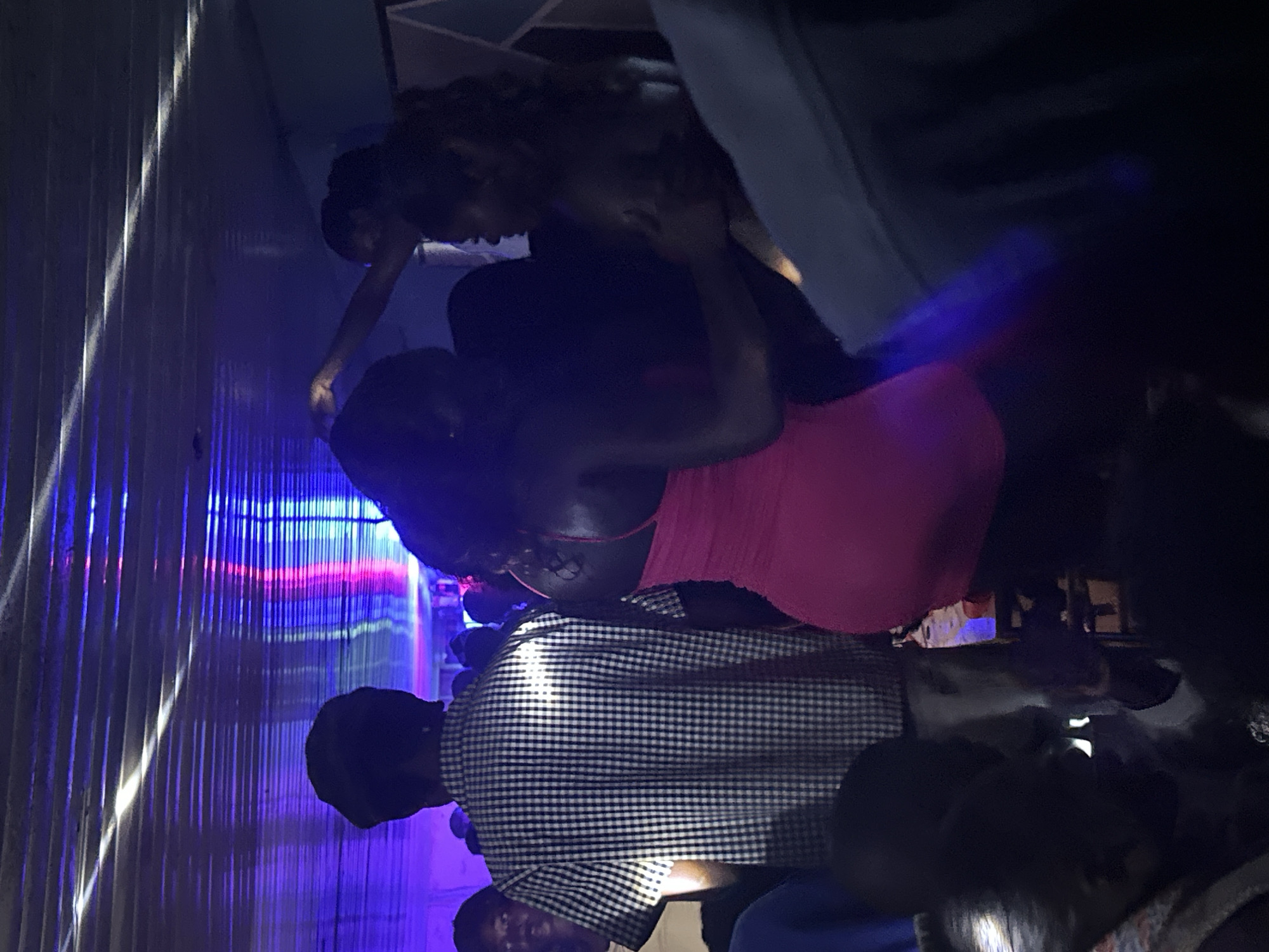 In Wumat, Nigeria, a dimly lit indoor space with a corrugated metal ceiling is illuminated by colored stage lights, casting blue and pink hues. Several individuals are visible, mostly obscured by darkness or the angles of the shot. In the foreground left, a person's head is partially visible, facing down. To their right, a person wearing a black and white checkered short-sleeved shirt is seen from the back, with a dark cap on their head. In the center, a woman in a bright pink, form-fitting dress is embraced by another individual whose face is not clearly visible. To the right of them, another person's face is partially visible in profile, looking away. The scene appears to be a gathering or event, possibly a party or nightclub, characterized by low lighting and dancing or socializing.