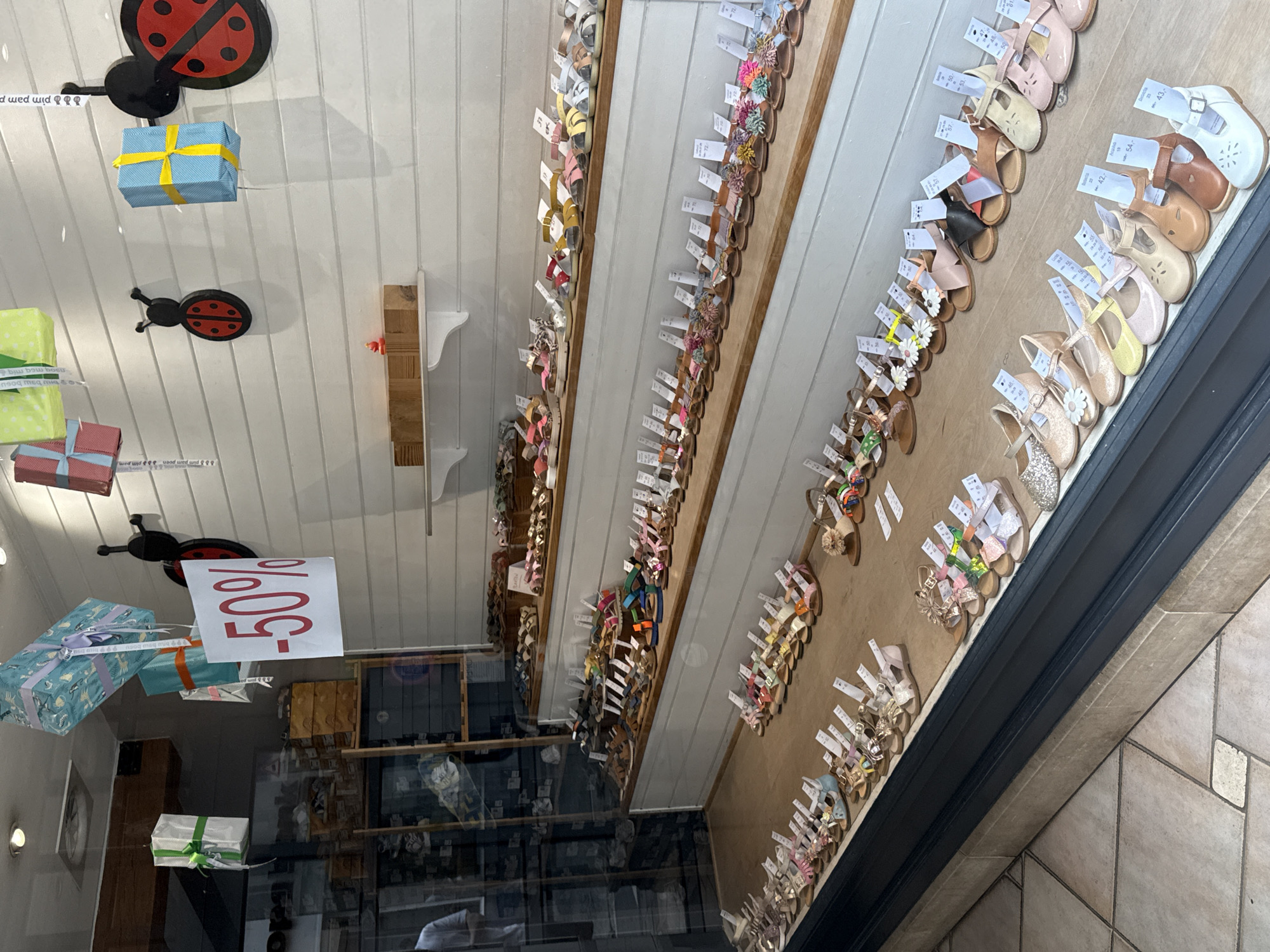 This is a medium shot, taken from a slightly elevated, exterior perspective, of a shop window in Bruges, Belgium. The shop appears to be a boutique selling children's shoes. The window displays rows of colorful sandals and closed-toe shoes, many with price tags. The shoes are arranged neatly on wooden shelves that are painted white. In the background, on the white paneled wall of the shop interior, are several decorative gift-wrapped boxes and two ladybug-themed decorations. A " -50%" sign is prominently displayed in the window. The floor in front of the display is tiled. No people are directly visible in the shop window, though a partial reflection of a person can be seen in the upper left portion of the window.