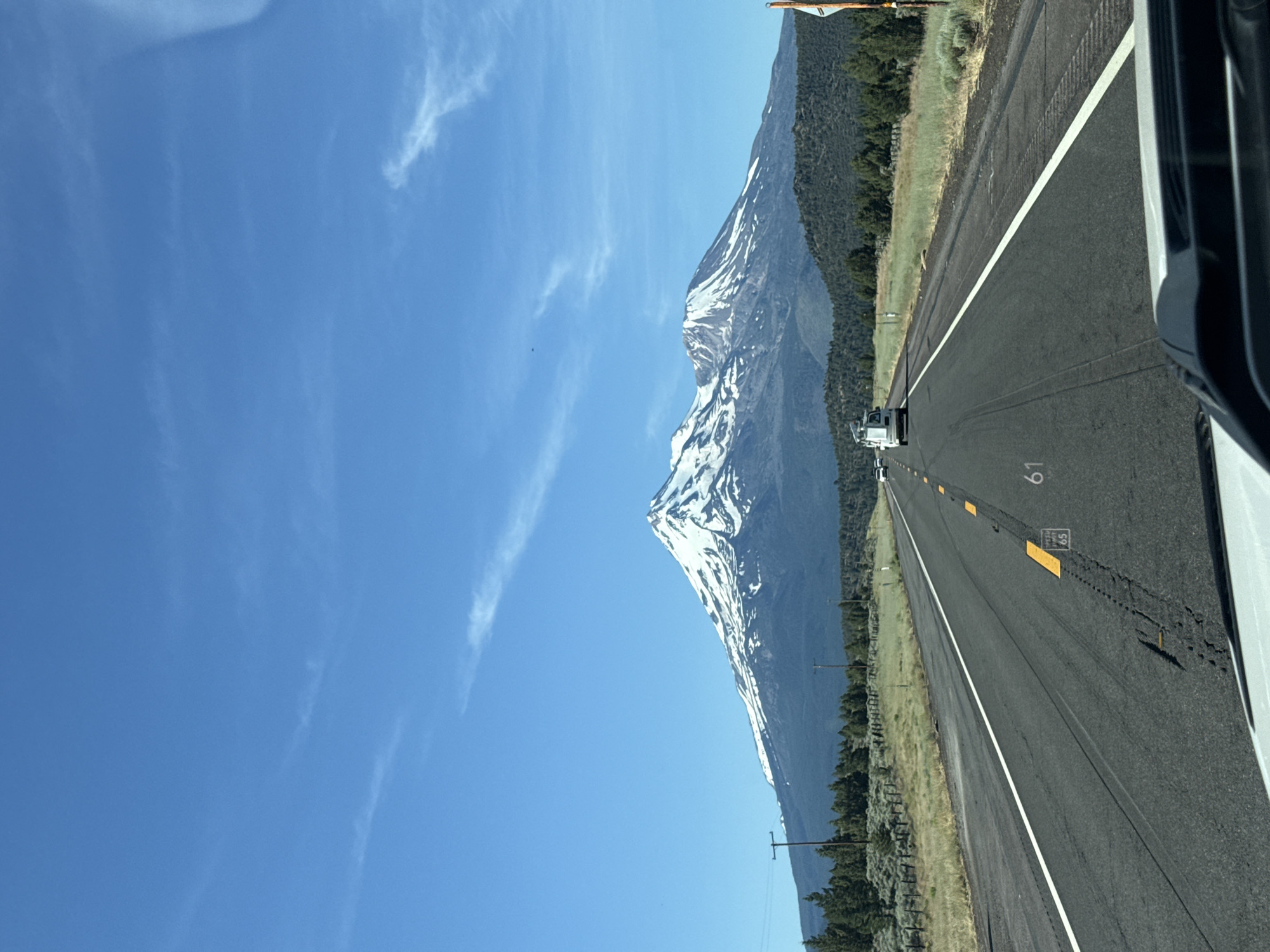 The image depicts a view from a vehicle traveling on a highway. The highway runs parallel to a large, snow-capped mountain. The mountain is covered in patches of snow, contrasting with the dark rock and green vegetation on its lower slopes. A dense forest of trees flanks the mountain and extends towards the roadside. The sky is a clear, bright blue with a few wispy white clouds.
The road appears to be a multi-lane highway with a white dividing line. Several yellow markers are visible along the side of the road, some with numbers. One marker clearly shows the number "65", and another further down the road has "61" visible. A white truck is seen in the distance, traveling in the same direction as the viewer. The right side of the image shows the interior of a vehicle, suggesting the photo was taken from the passenger seat or driver's side.
The scene is set during the daytime, likely late morning or early afternoon, given the bright sunlight and clear sky. The weather appears to be pleasant, with sunshine and a lack of precipitation. The presence of snow on the mountain suggests a location at a higher altitude or a cooler climate. The highway and the surrounding landscape indicate a rural or semi-rural setting. The overall impression is one of a scenic drive through a mountainous region. There is no indication of people in the image, apart from the implied presence of the photographer and any occupants of the vehicle.