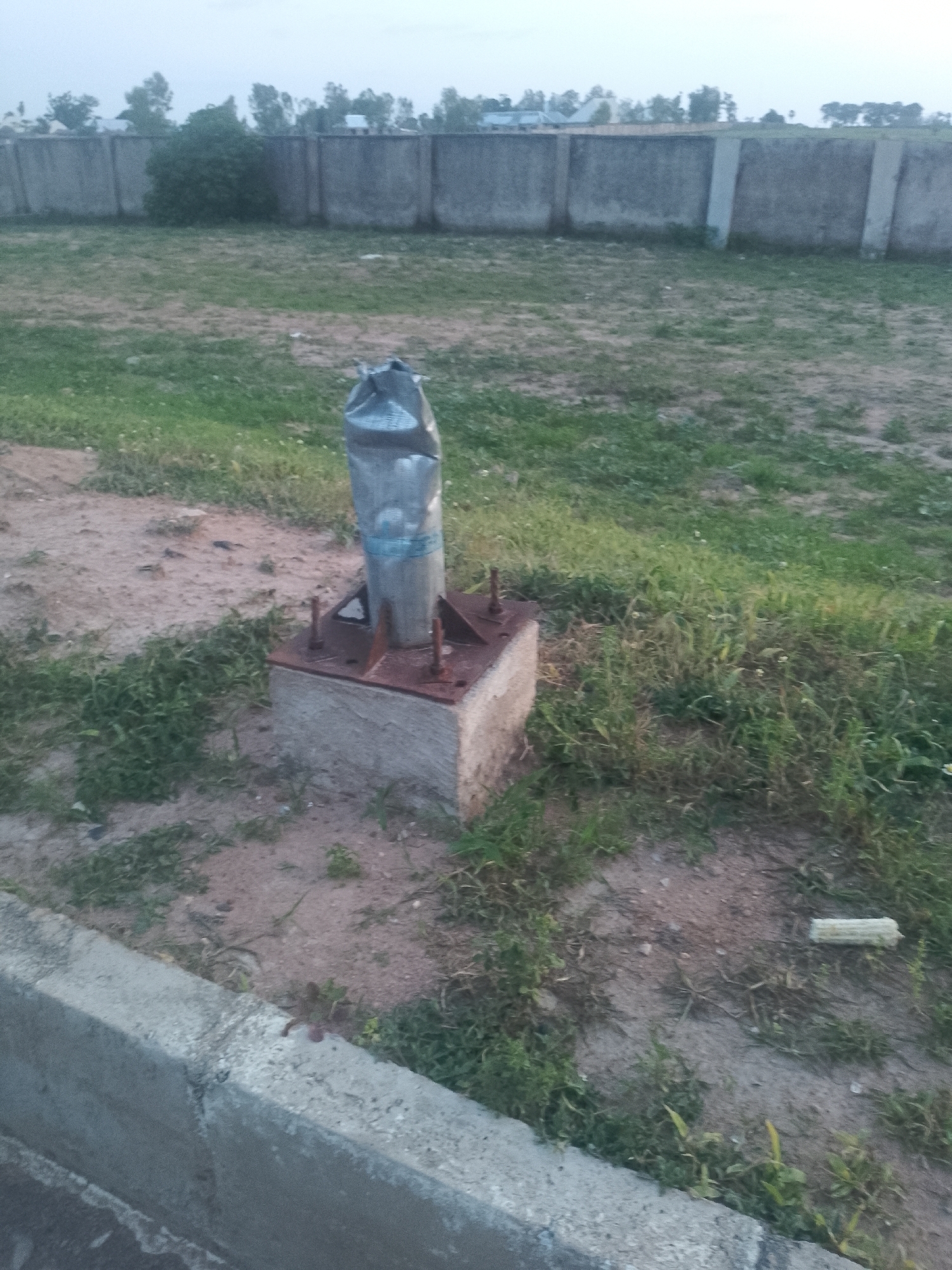 In an outdoor, grassy area with a concrete wall in the background, a cylindrical object wrapped in plastic is mounted on a concrete base. The object appears to be metallic and is secured to the base by bolts. A piece of corn cob lies on the ground to the right of the object. The scene is viewed from a low angle, with a concrete curb in the foreground.