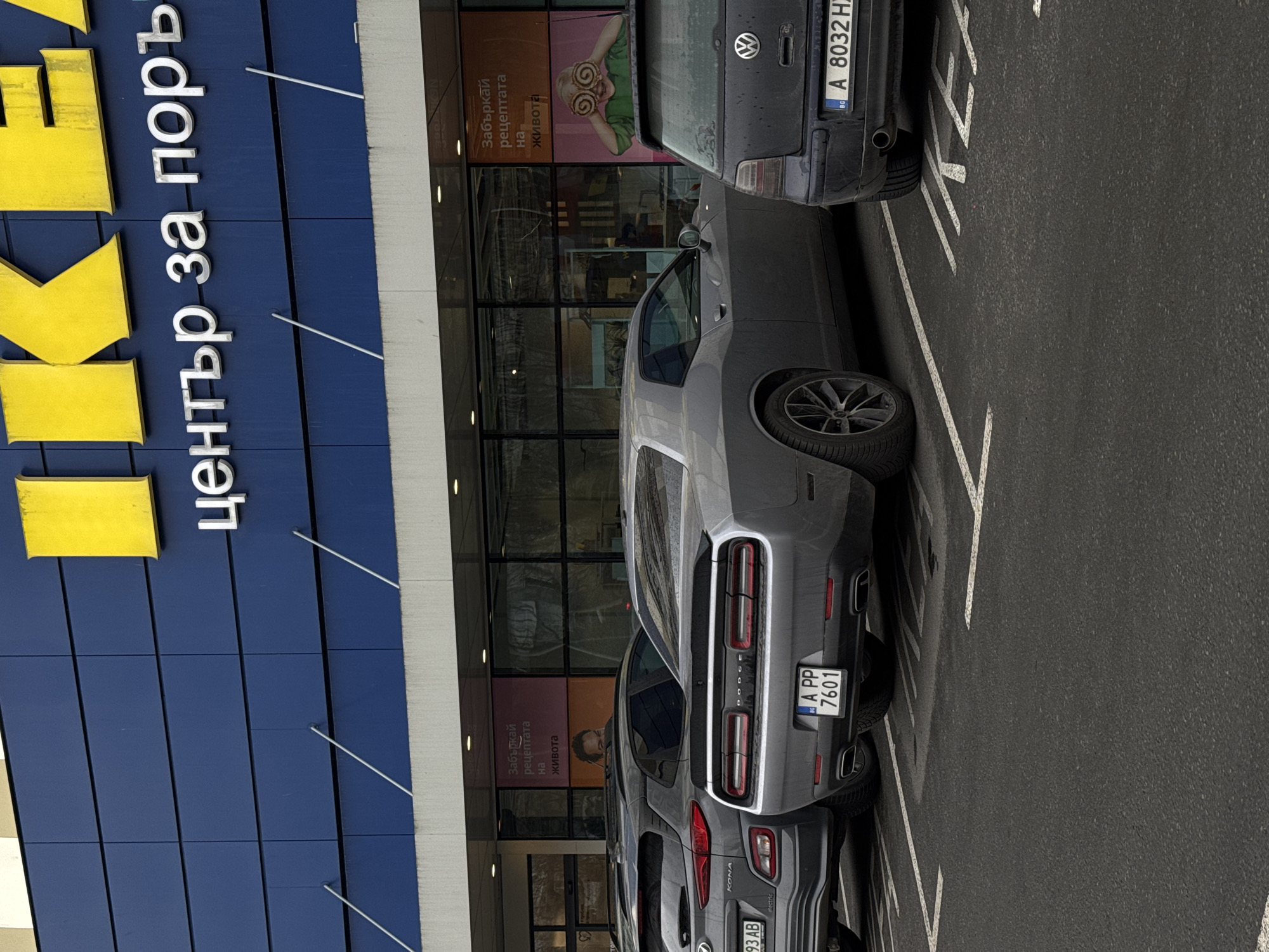 The image depicts the exterior of an IKEA store in Burgas, Bulgaria. The building is blue with the distinctive yellow IKEA logo and the text "център за поръчки" in Bulgarian. In front of the store are parked cars, including a gray Dodge Challenger with license plate "A PP 7601" and a dark gray Volkswagen with license plate "A 8032 HZ". There are advertisements visible in the windows, including one showing a person with food over their eyes and the text "Забъркай рецептата на живота" (Mix the recipe of life). The parking lot is paved and marked with white lines.
