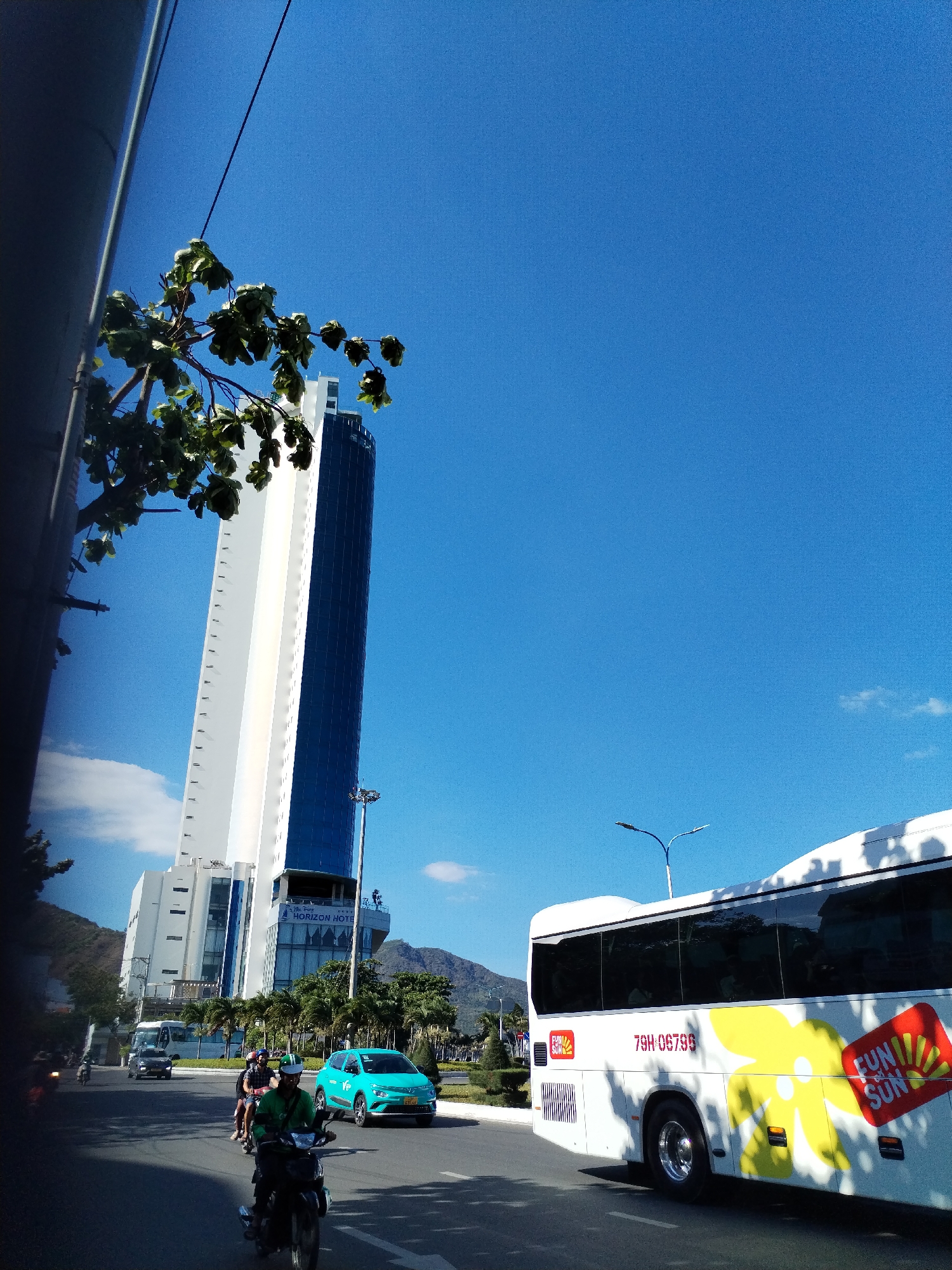 The image depicts a street scene in Nha Trang, Vietnam, specifically in the Bac Nha Trang Ward. A tall building, identified as the Horizon Hotel, dominates the background, with its white and blue facade. In the foreground, a white tour bus with a distinctive "Fun Sun" logo and license plate "79H-06796" is partially visible. A street runs through the middle ground with traffic including motorbikes and a turquoise car. A man on a motorbike is directly in the foreground. The sky is clear and blue. Mountains can be seen in the distant background. A tree branch with green leaves is visible on the left side. A streetlight is present near the Horizon Hotel.